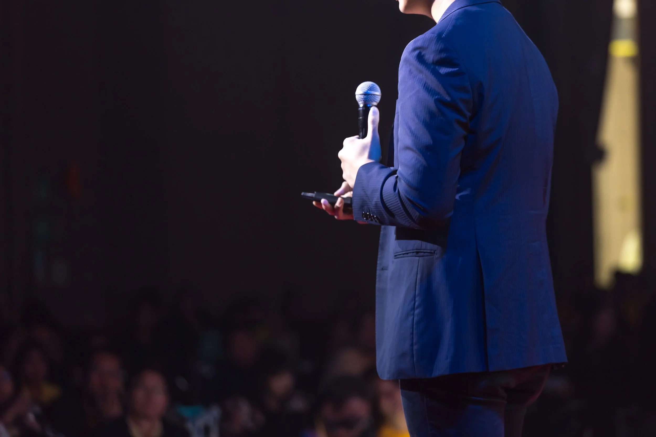 Person in a blue suit holding a microphone and a phone on stage in front of an audience.