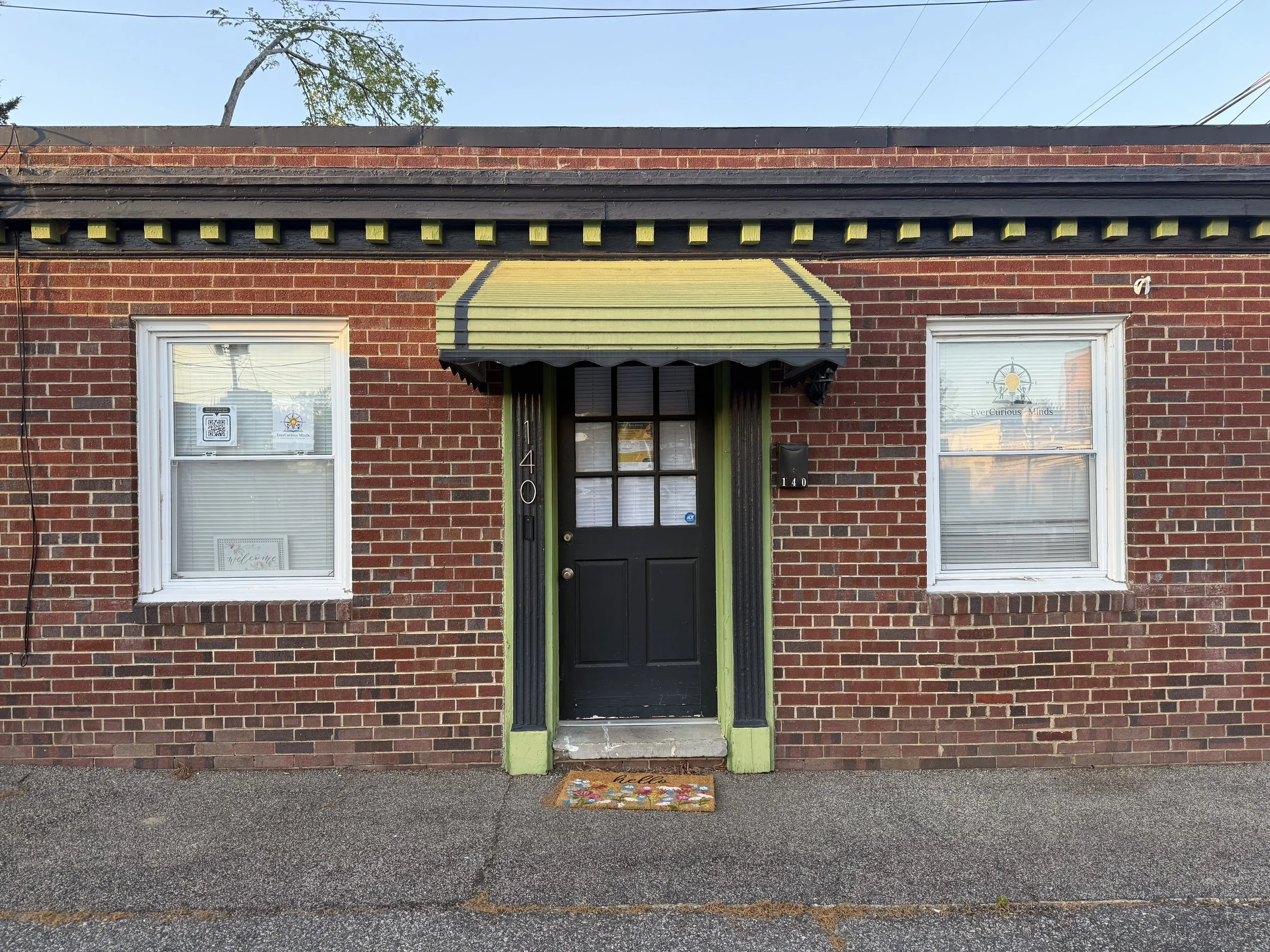 Front brick building with black door, two windows, and green trim. The windows have decorations and signs, with a welcome mat in front of the door.