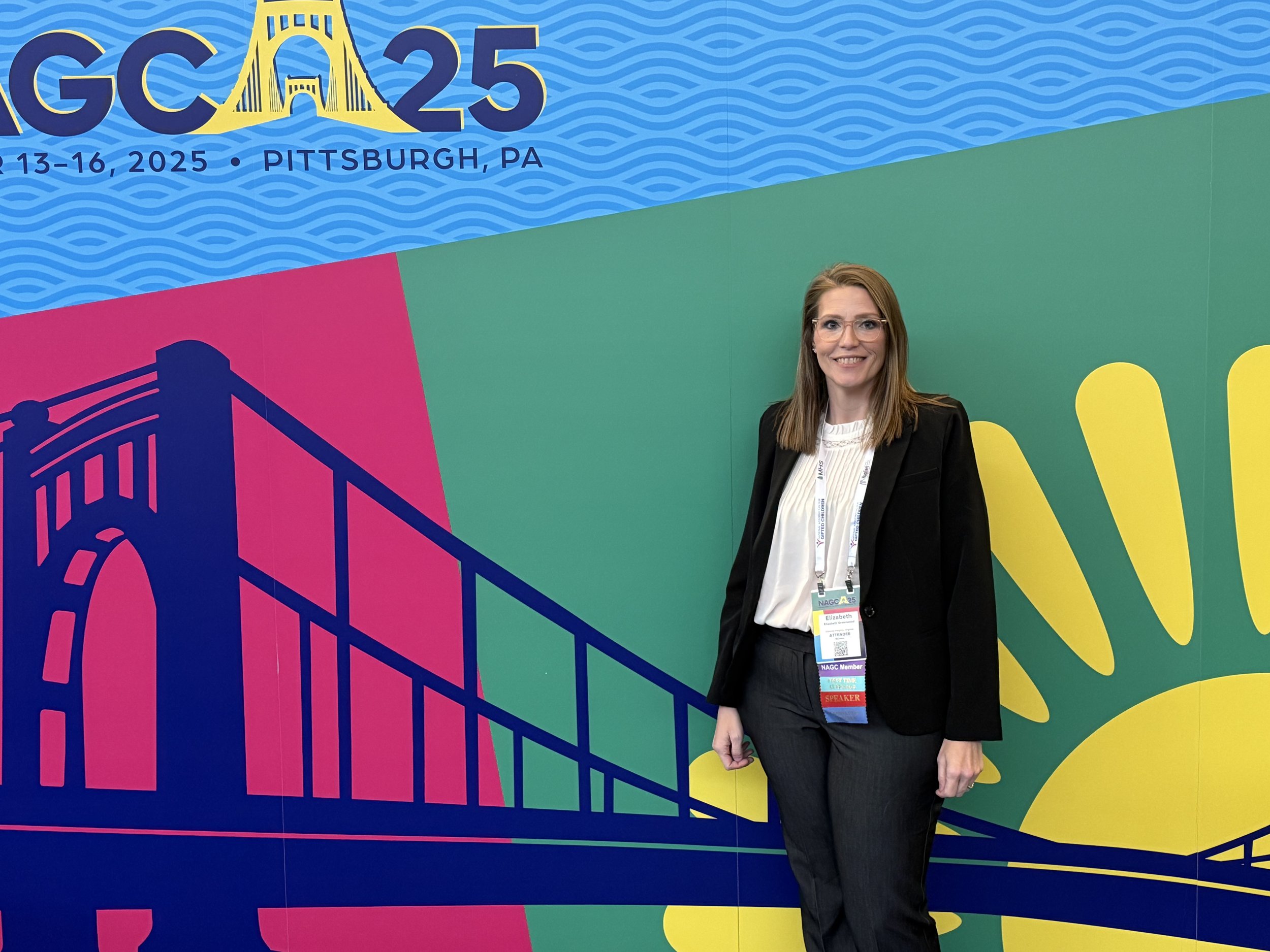 A woman standing in front of a colorful conference backdrop with a stylized bridge, wavy blue pattern, and event details, smiling and wearing a black blazer and pants with conference badges.