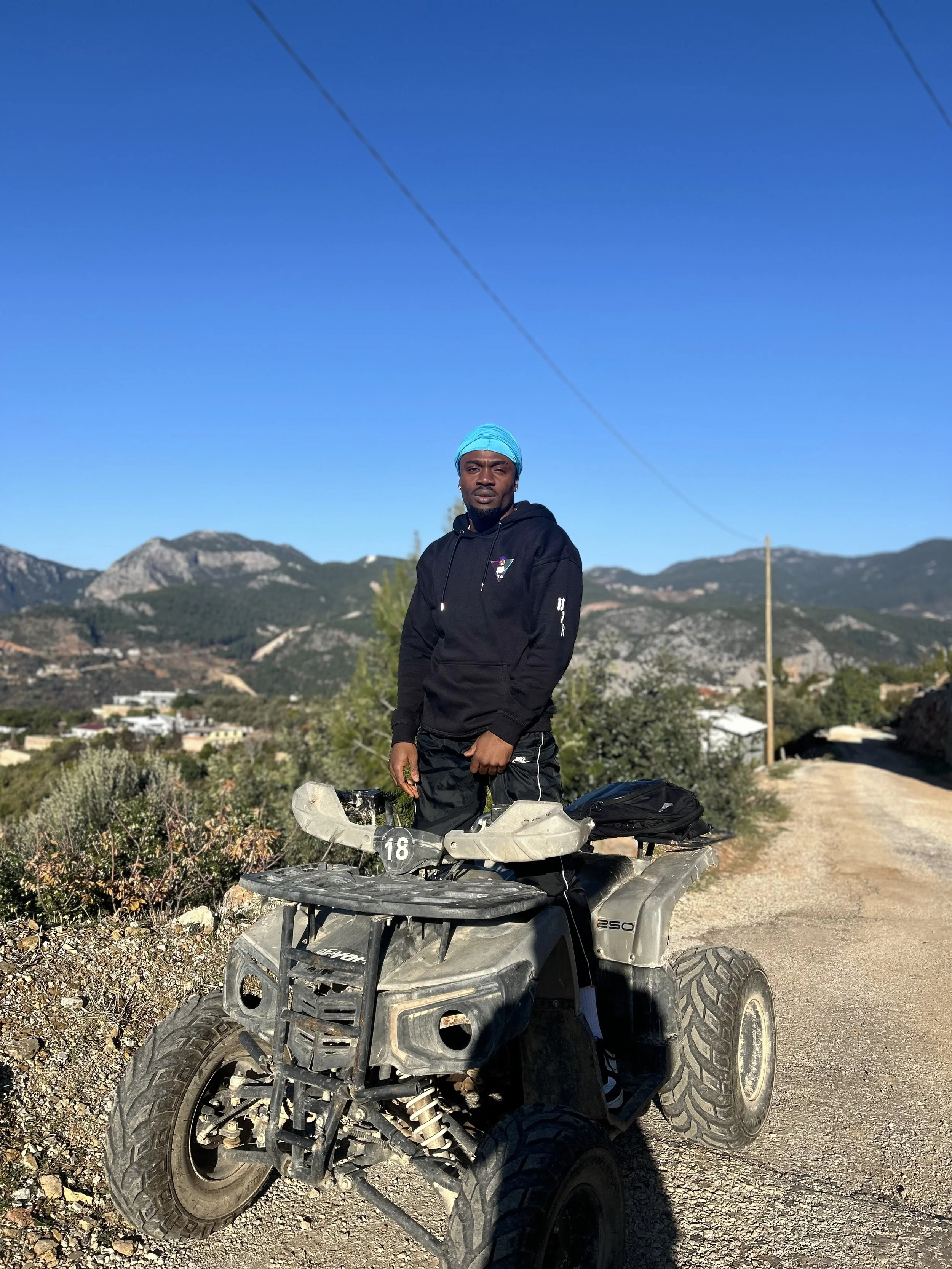 Mann steht auf einem ATV in einer ländlichen bergigen Landschaft bei sonnigem Wetter, trägt schwarze Kleidung und einen blauen Haarschal.