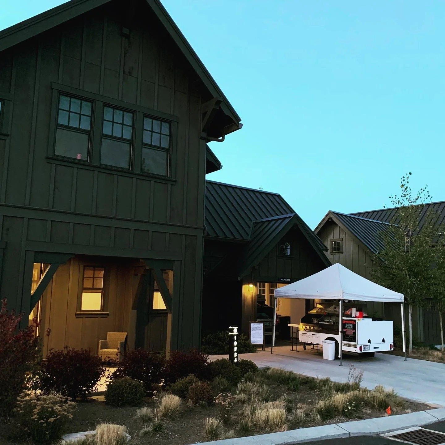 A residential building with green siding and a metal roof, featuring large windows. In front, there is a white tent set up over Bruneau's food cart, with lights illuminating the scene at dusk. There are bushes and plants in the foreground.