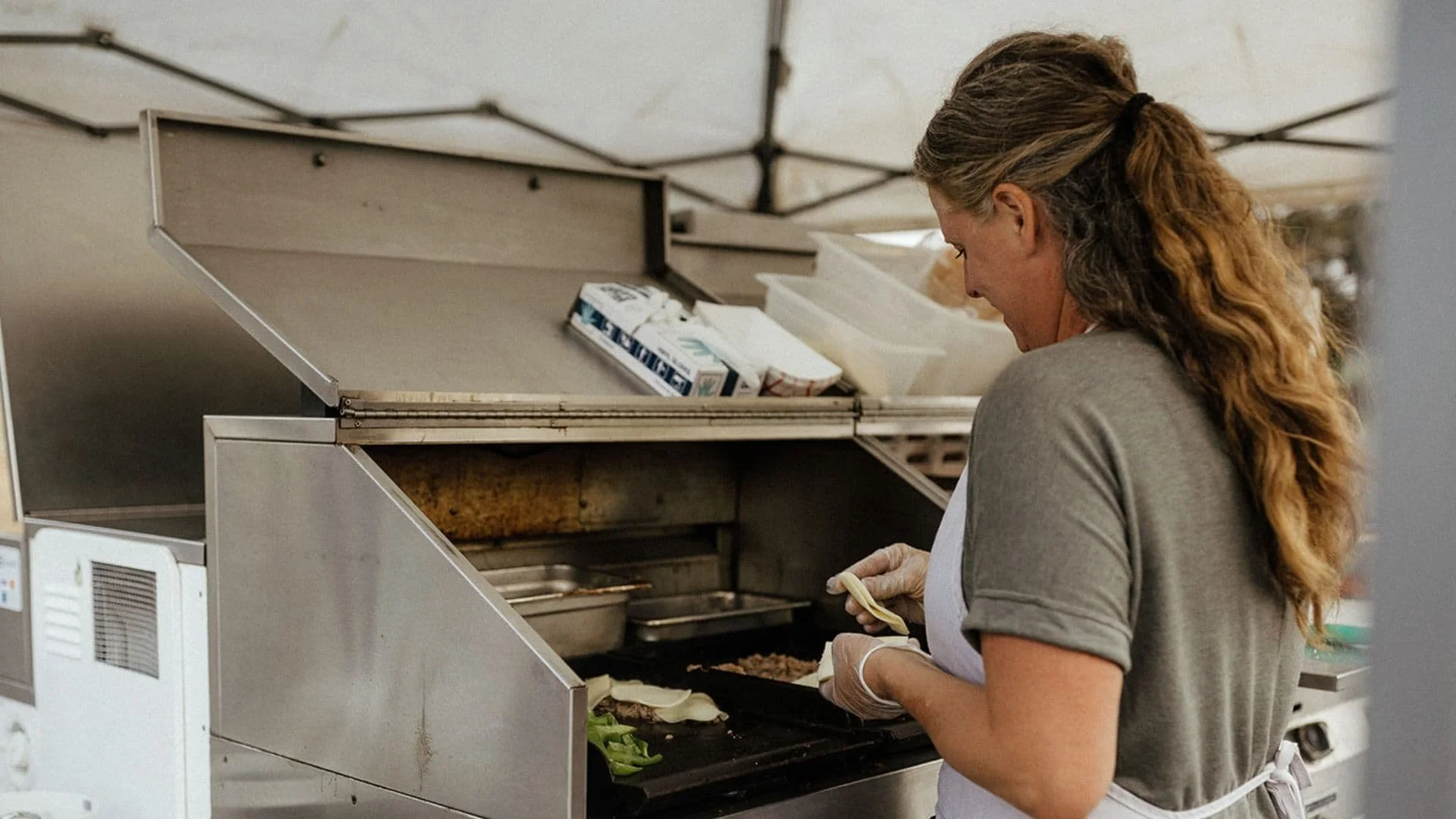 A woman with long, curly hair preparing food at an open-air food cart, wearing a gray shirt, a white apron, and gloves, with food items and kitchen equipment around her.