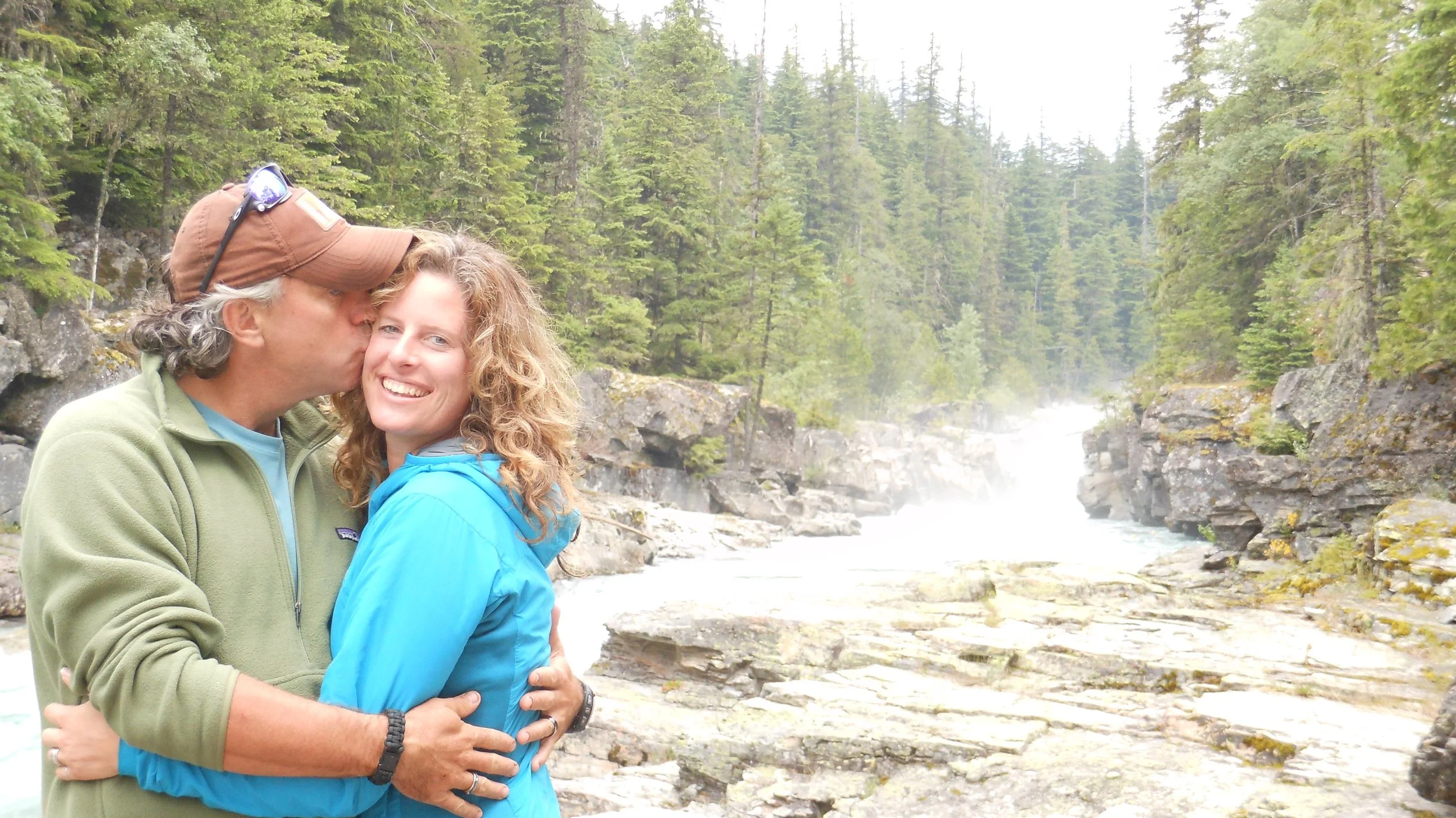 A man kisses a woman on the cheek in an outdoor setting with a river and trees in the background