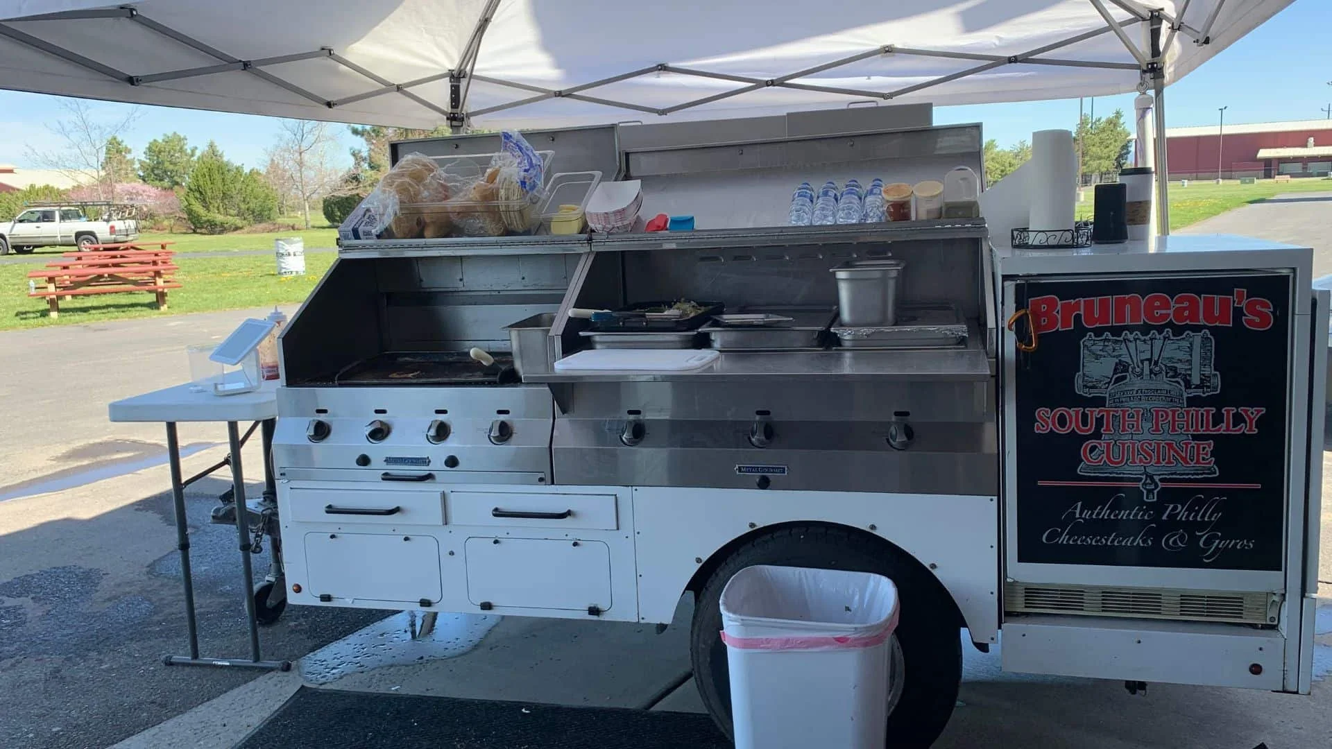 Food truck with a sign reading 'Bruneau's South Philly Cuisine, Authentic Philly Cheesesteaks & Gyros' parked outdoors under a canopy, with supplies and cooking equipment visible on the counter.