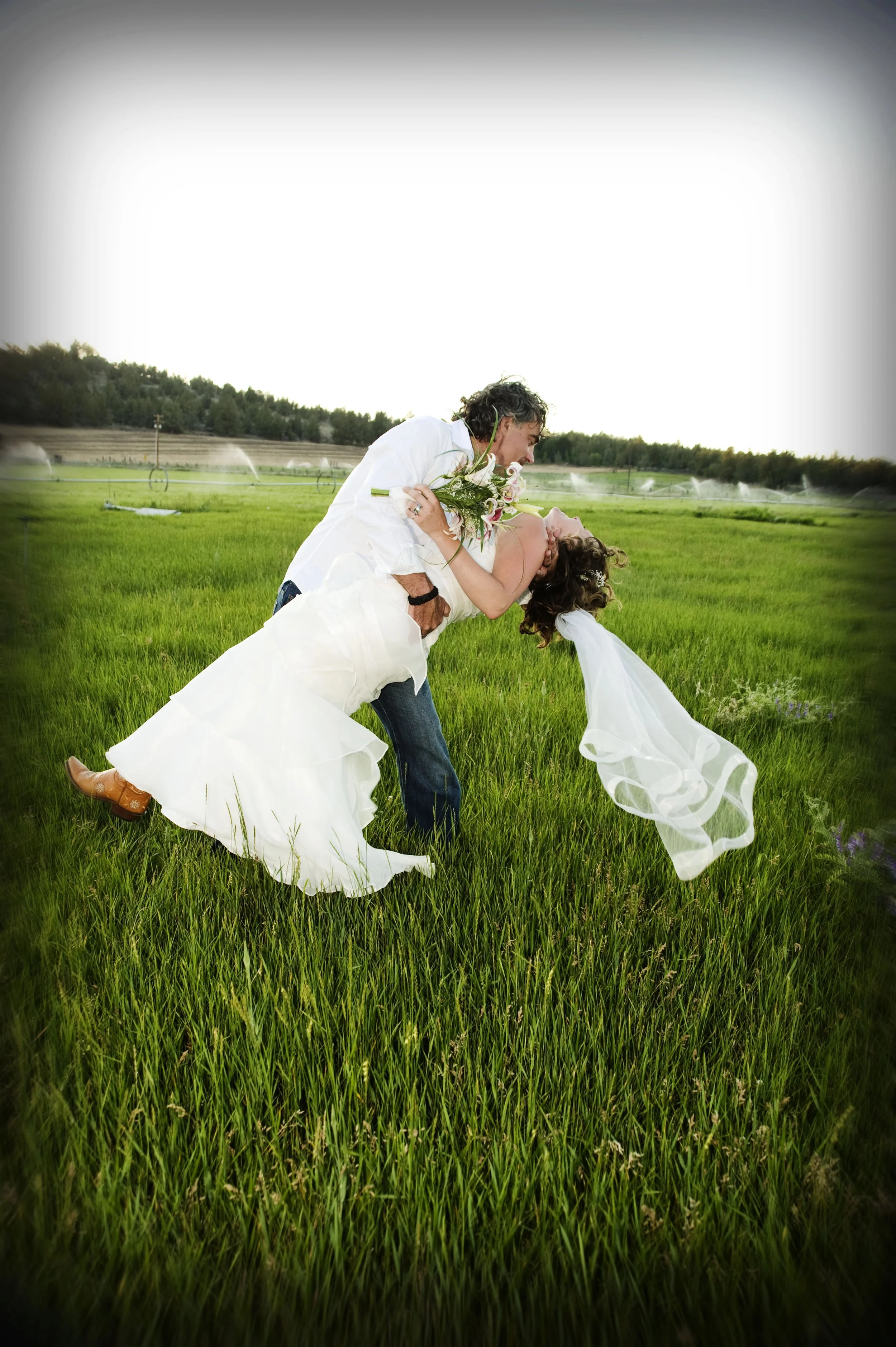 A couple dancing in a green field, the man dipping the woman while she holds a bouquet, on a bright day.