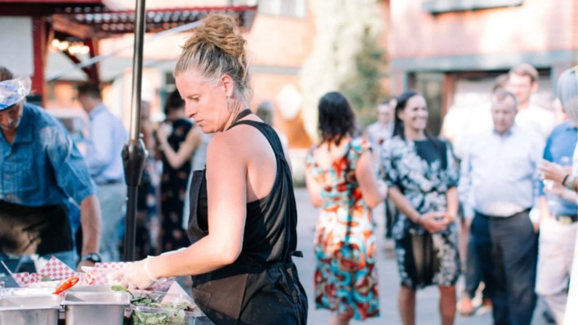 Woman serving food at an outdoor wedding with people mingling and talking in the background.