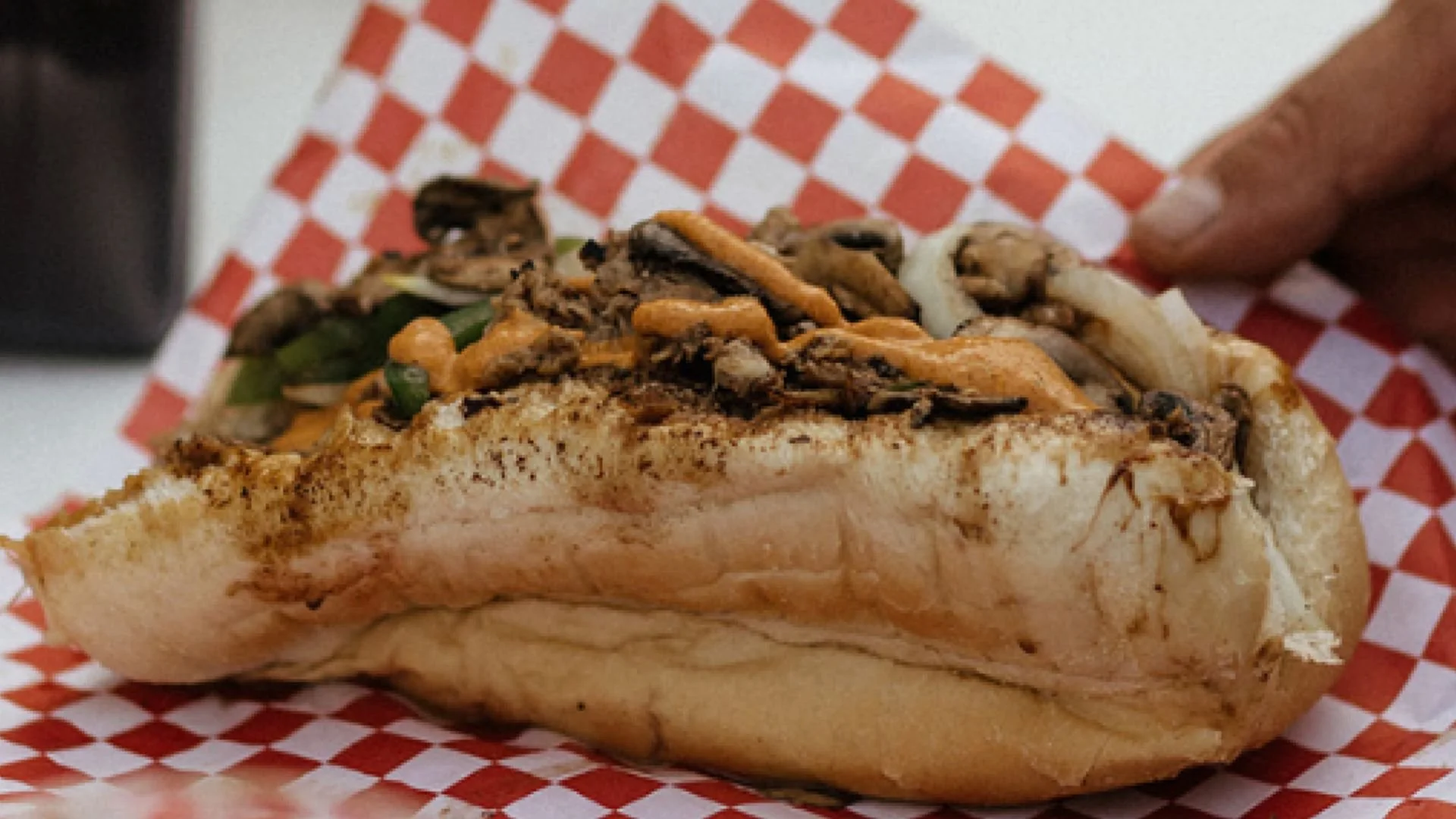 Authentic Philly Cheesesteak with chip-rib eye steak, sautéed mushrooms, onions, and green peppers on an Amorosa-style roll, placed on red and white checkered paper.
