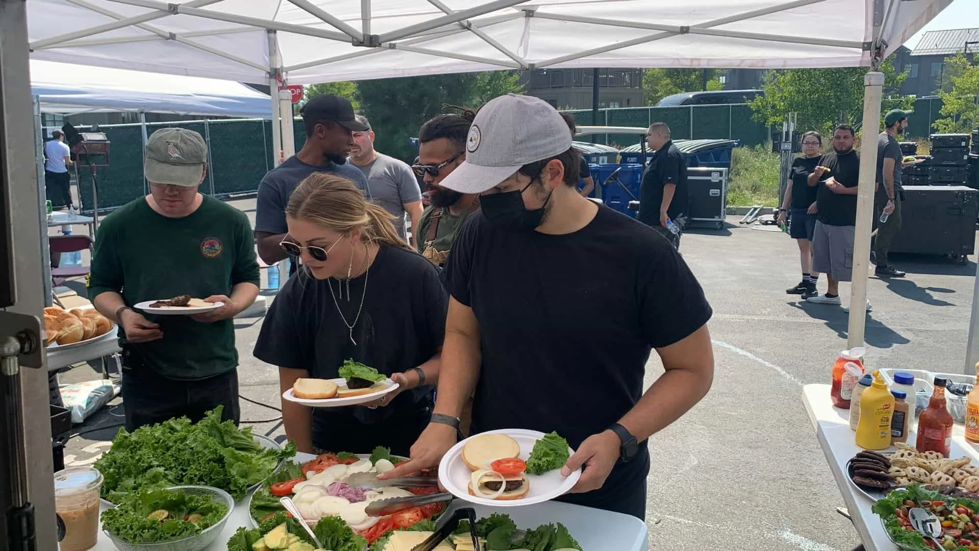 People in line at an outdoor food cart and buffet line, serving and preparing salads, sandwiches, and condiments under a white canopy tent on a sunny day.