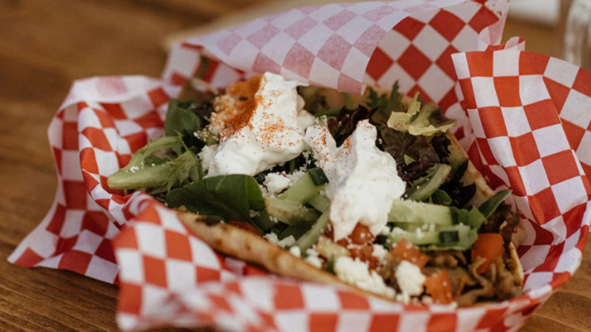 Bruneau's Signature sandwich, a Traditional Greek Gyro is in a basked line red and white checkered paper.  A pita is filled with green lettuce, gyro meat, white tzatziki sauce, cucumbers, and tomatoes and sprinkled with red paprika.