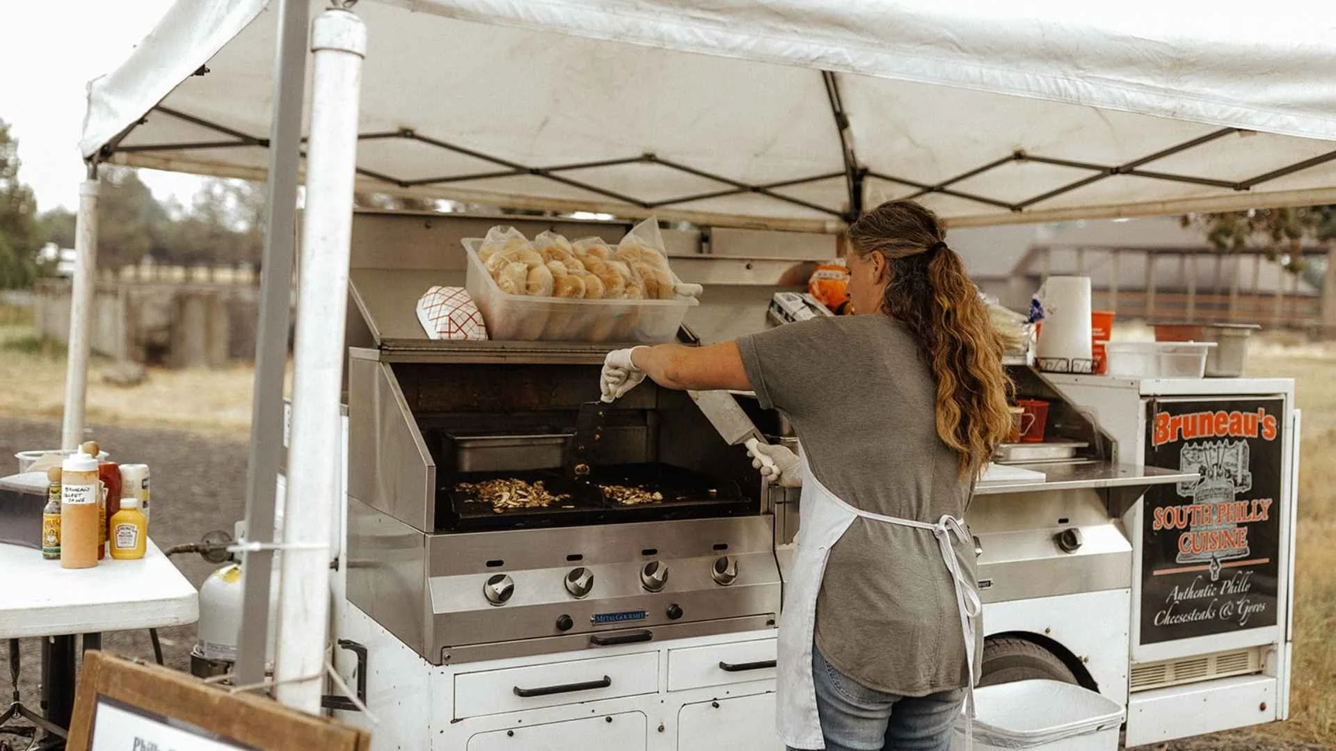 Woman grilling food at a food stand with a sign that says "Bruneau's South Philly Cuisine" and a tray of baked goods nearby.