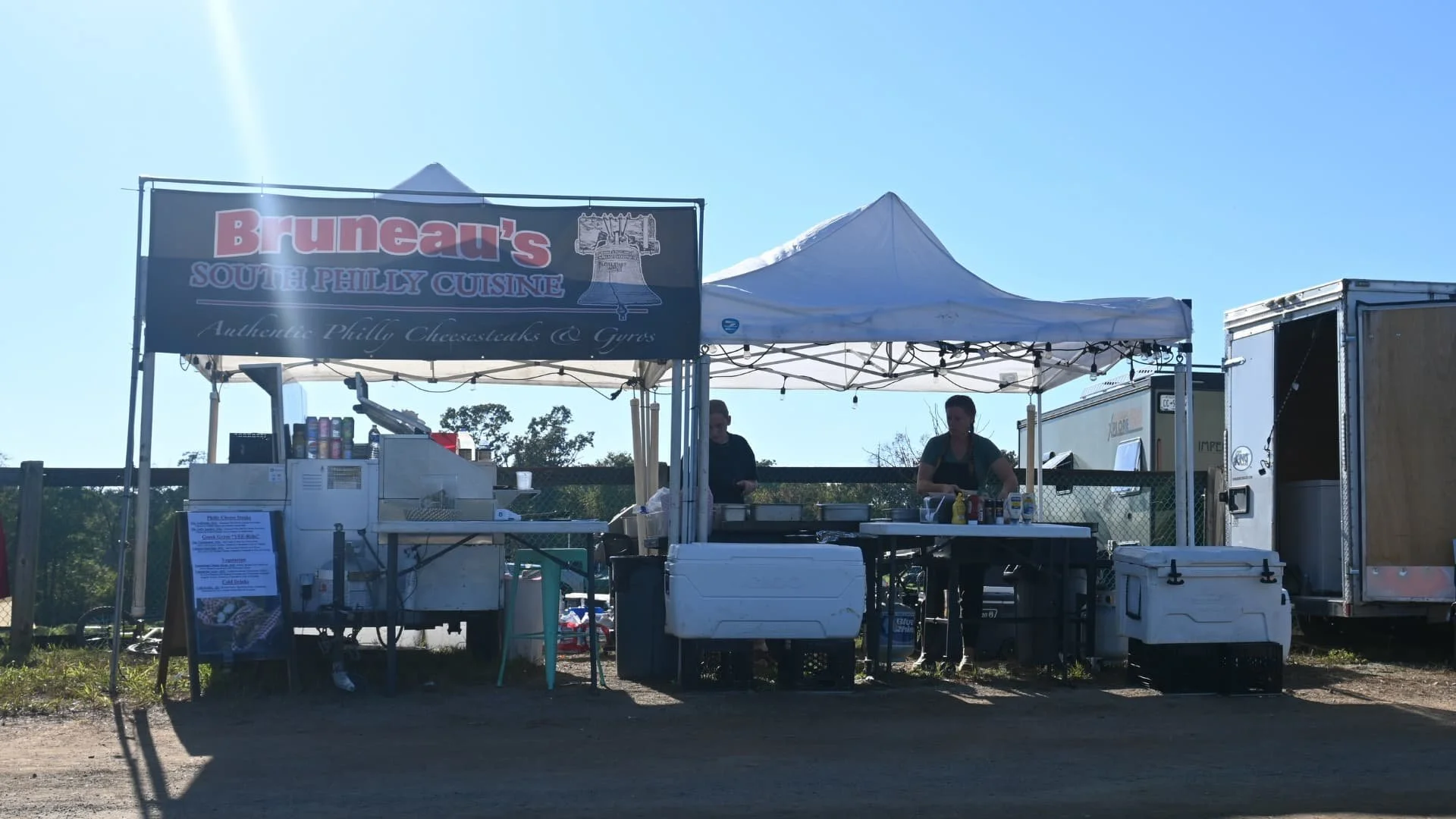 A street food stand with a sign reading 'Bruneau's South Philly Cuisine' selling Philly cheesesteaks and gyros, with two people preparing food under a white tent on a sunny day.
