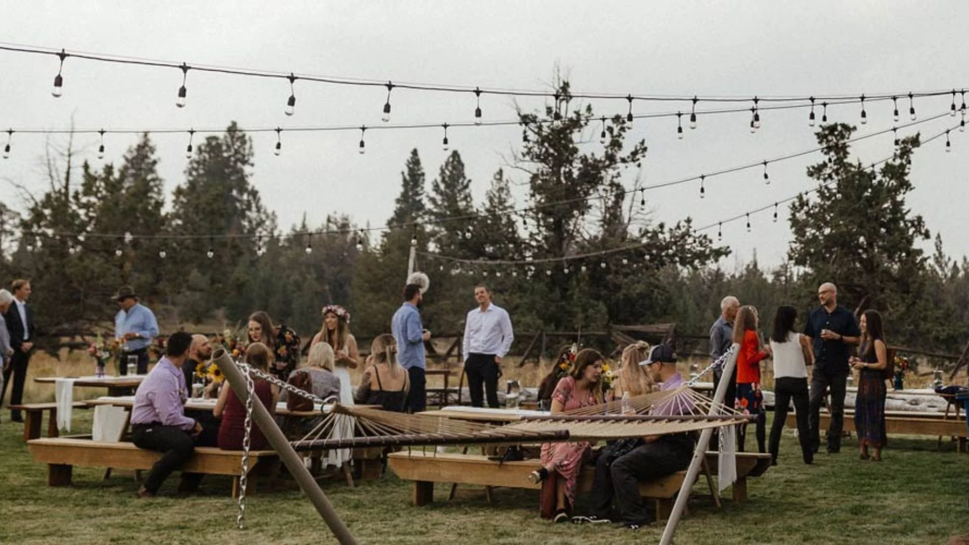 People socializing at an outdoor wedding in a grassy field with string lights overhead and trees in the background, some seated at tables and others standing and talking.