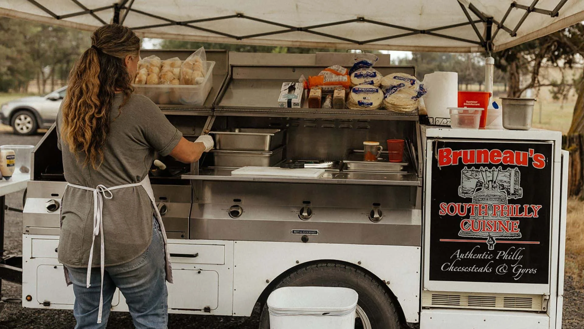 A woman preparing food at an outdoor open-air food cart with the logo "Bruneau's South Philly Cheesesetaks", featuring various ingredients and supplies on the counter.