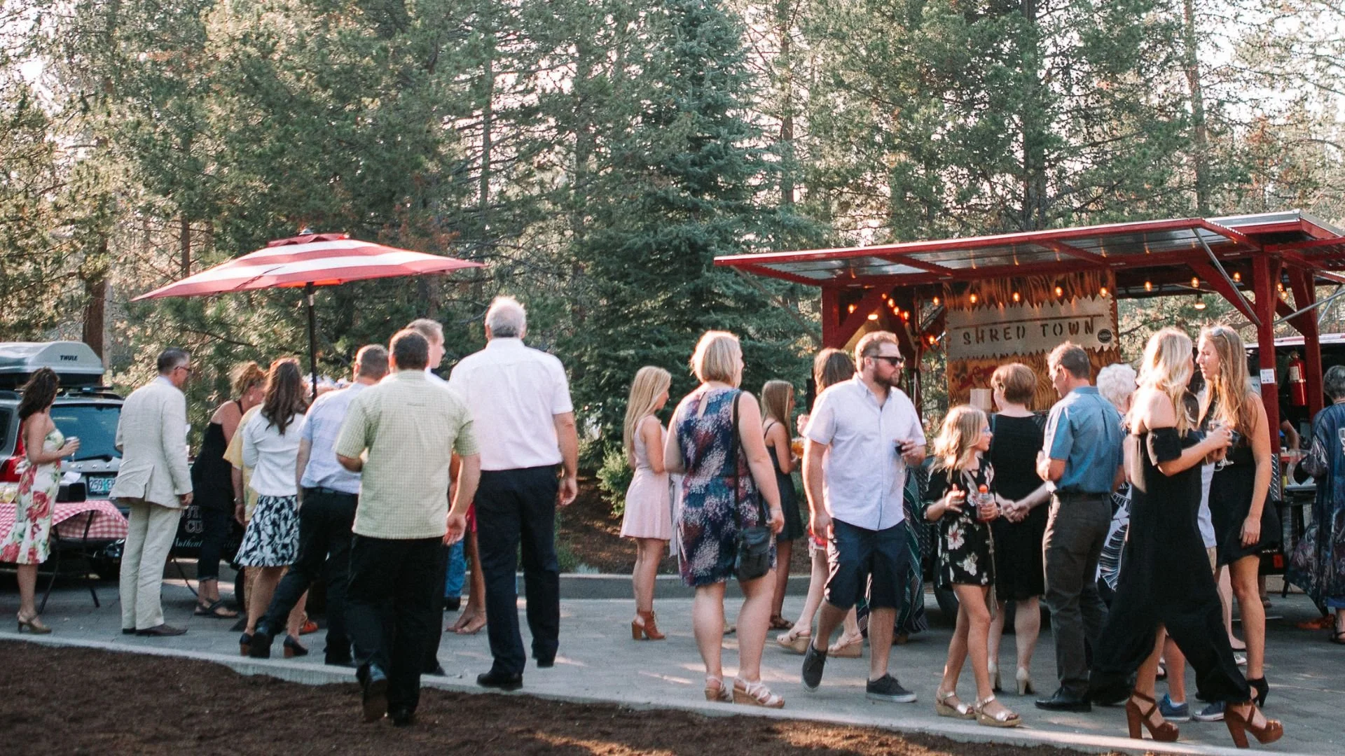 People dancing and socializing outdoors at a gathering with food trucks, trees, and string lights in the background.