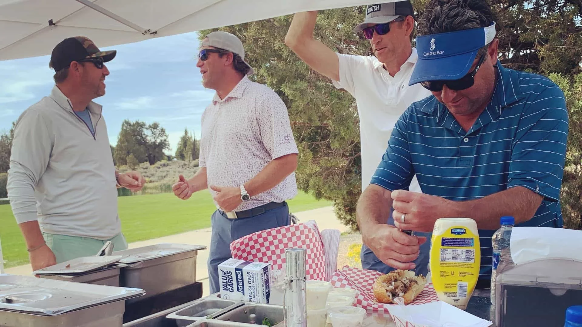 Four men are gathered at an open-air food cart on a golf course, two are talking, one is putting condiments on a sandwich, and one is relaxed with his hands on the umbrella above. 