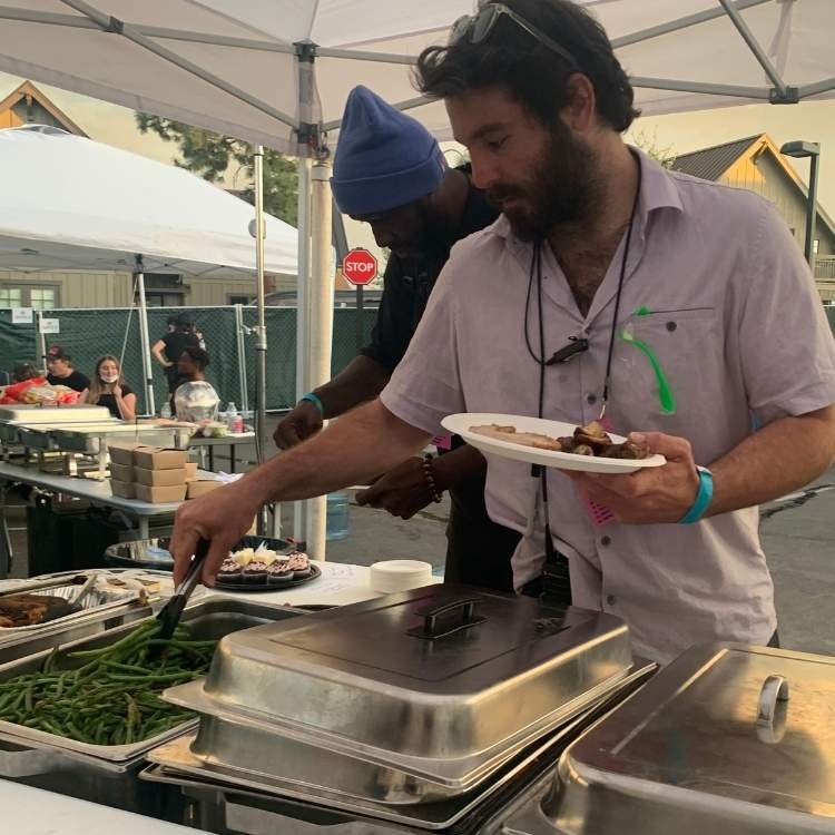People at an outdoor buffet line serving food from chafing dishes, with a man in a light shirt holding a plate of food in the foreground.