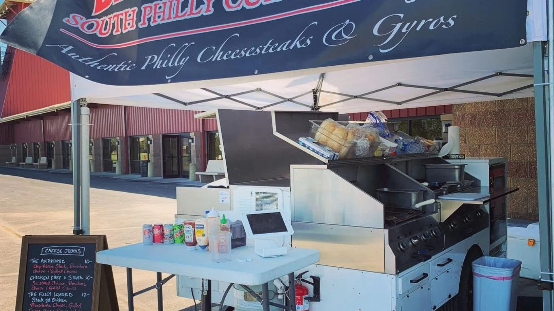 A food stand under a tent with a sign reading 'Bruneau's South Philly Cuisine' and a menu board listing Philly cheese steaks. The open-air food cart has bread, bottles of condiments, and a grill visible.