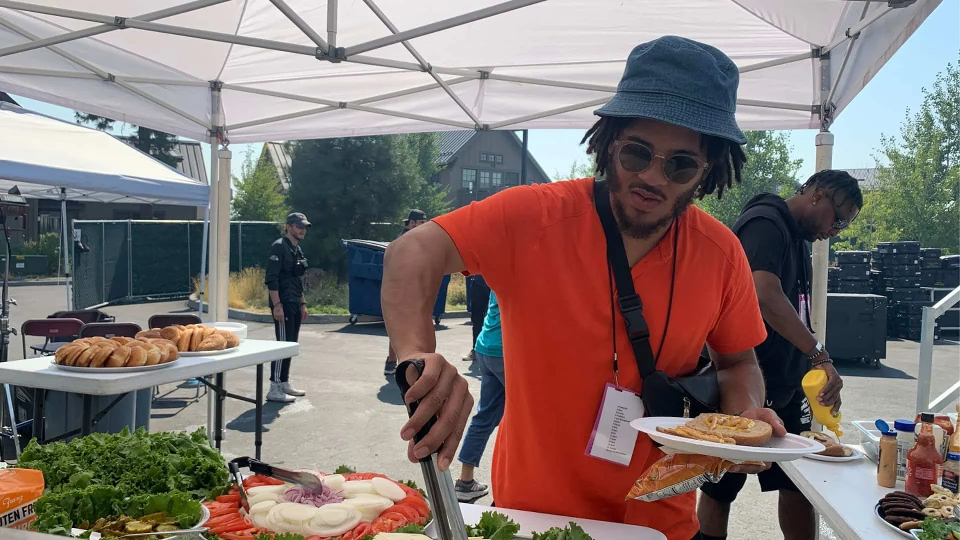 A man from a film crew in a bright orange t-shirt, wearing sunglasses and a blue bucket hat, is serving food at an outdoor event table. There are trays of food on the table, with a few other people in the background under a white canopy, 