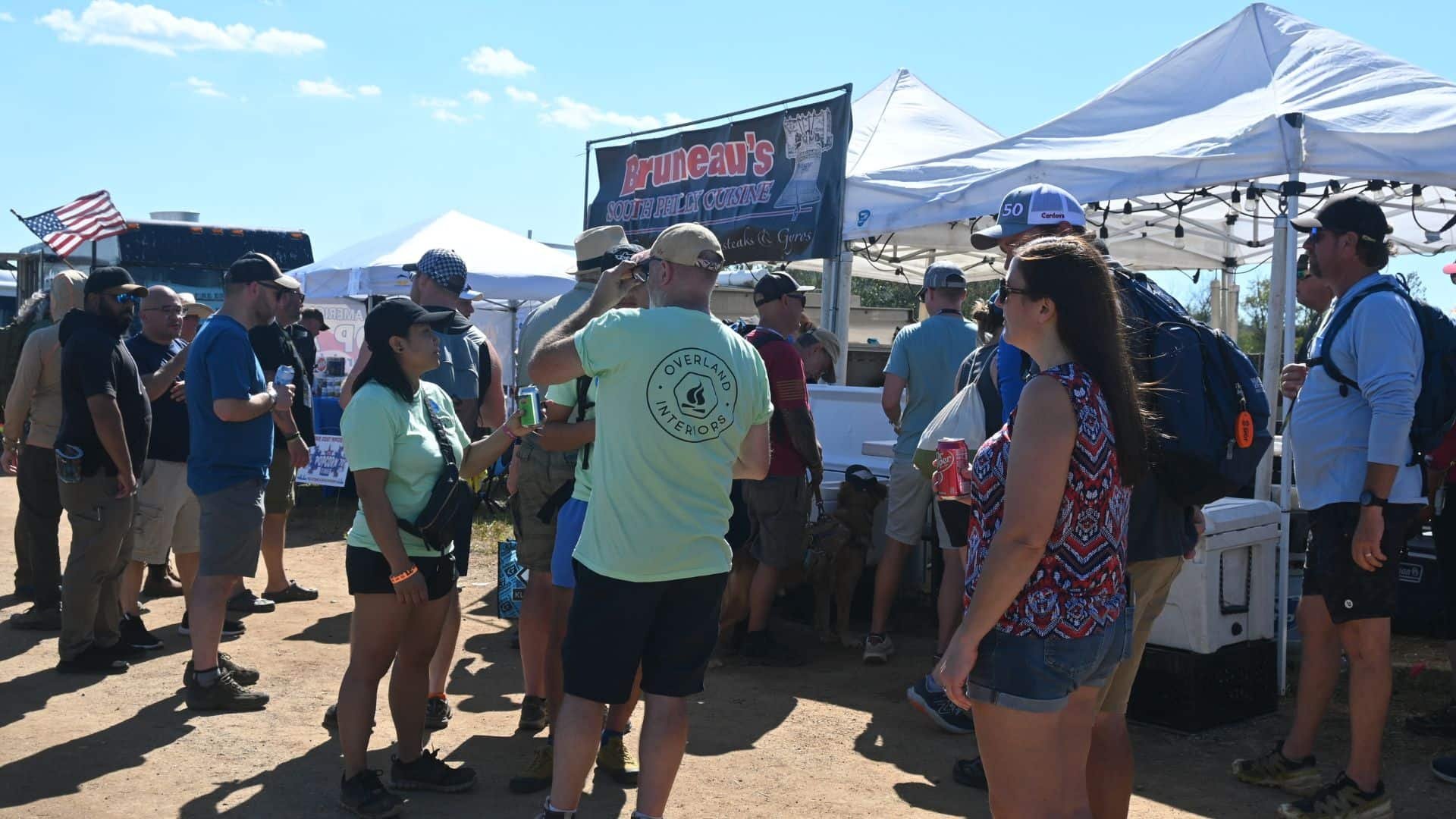 People gather around food stalls at an outdoor event on a sunny day, with tents and an American flag visible.