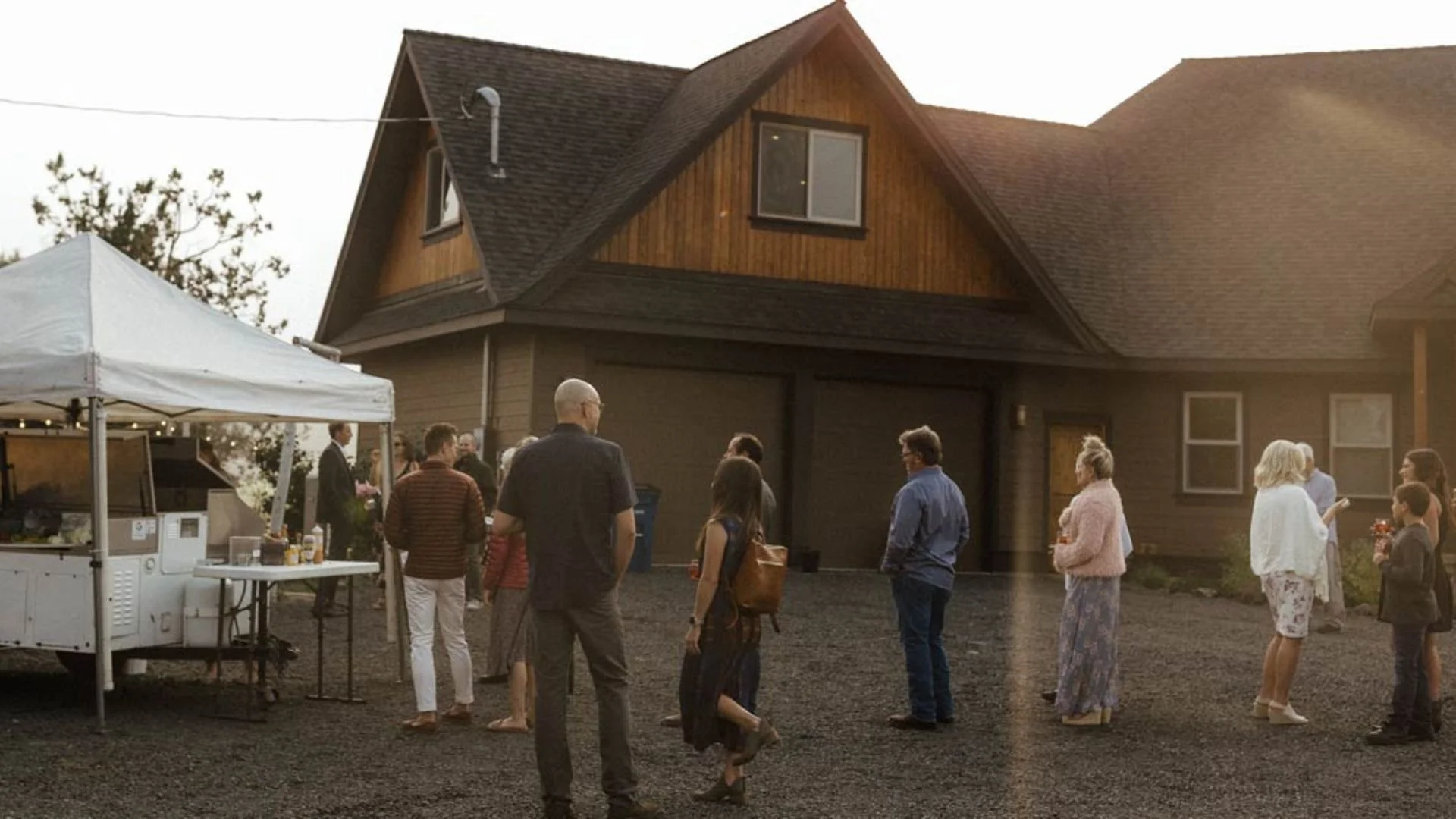 People gathered outside a house during a wedding are lined up to get food at an open-air food cart under a white canopy tent. 