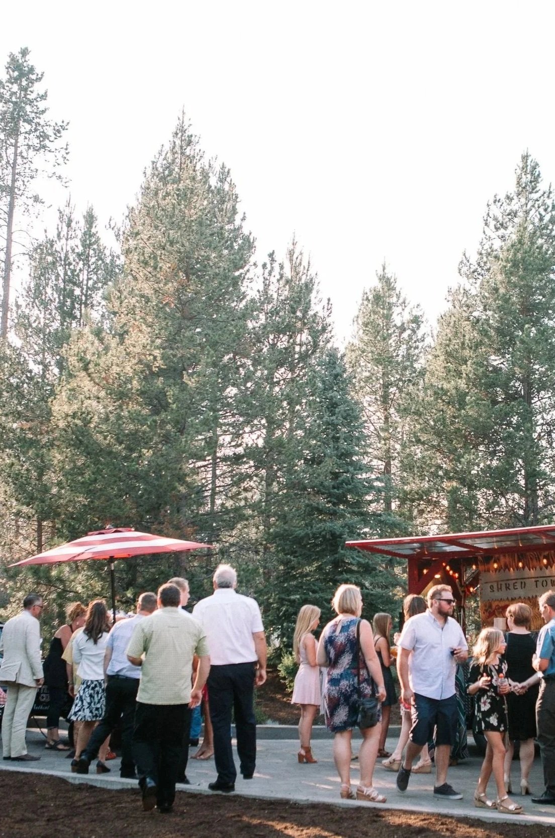 People gathered at an outdoor wedding, some dancing and socializing, while others are gathered ordering food in front of two food carts in the background.