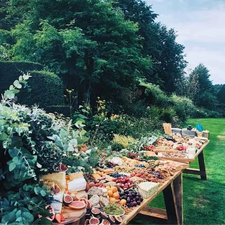 A long outdoor table overflowing with a variety of fresh fruits, vegetables, and snacks set in a lush garden with greenery and trees in the background.