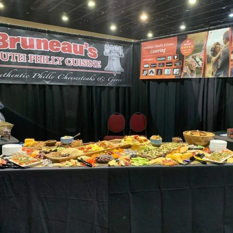 A food display table with various cheese and snack platters at an event, with black curtains and banners for Bruneau's South Philly Cuisine in the background.