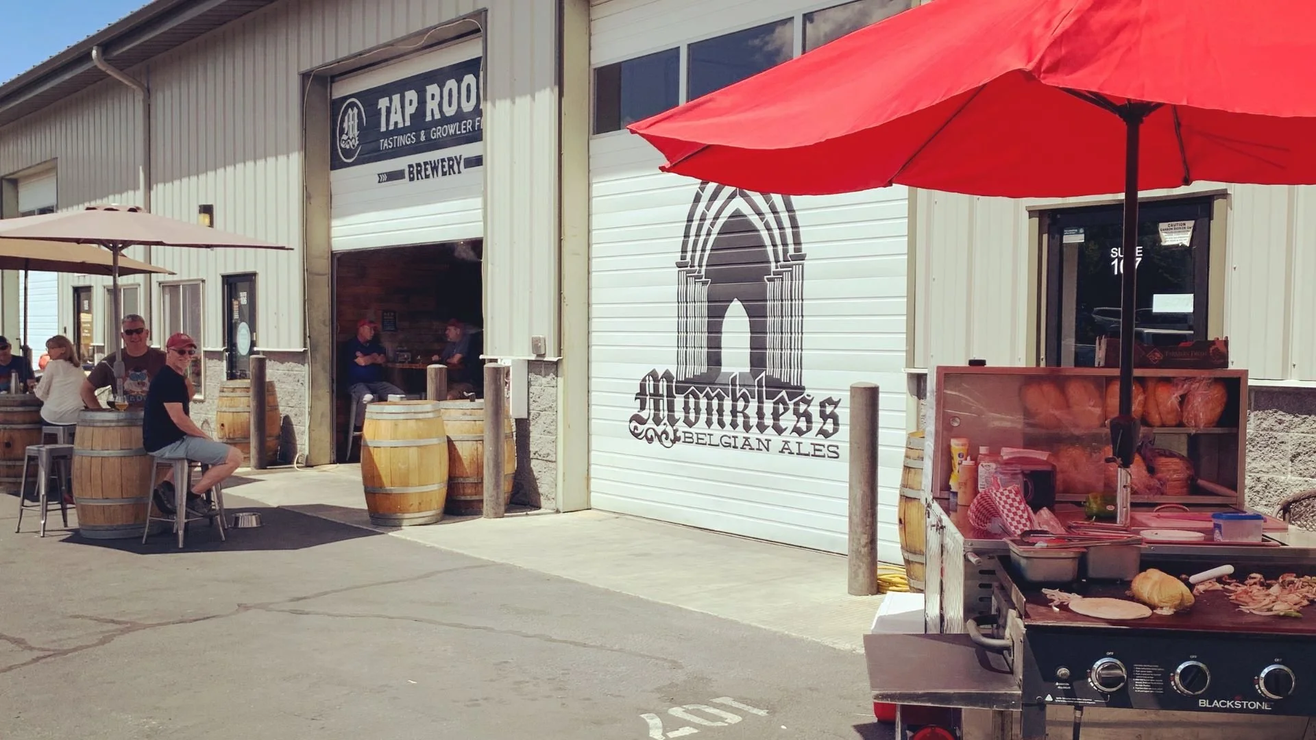 Outdoor patio of Monkless Belgian Ales, a brewery with customers sitting at high tables covered with barrels in front of a large open garage door, with a logo feat  In the foreground is Bruneau's South Philly Cuisine's original, open-air food cart.