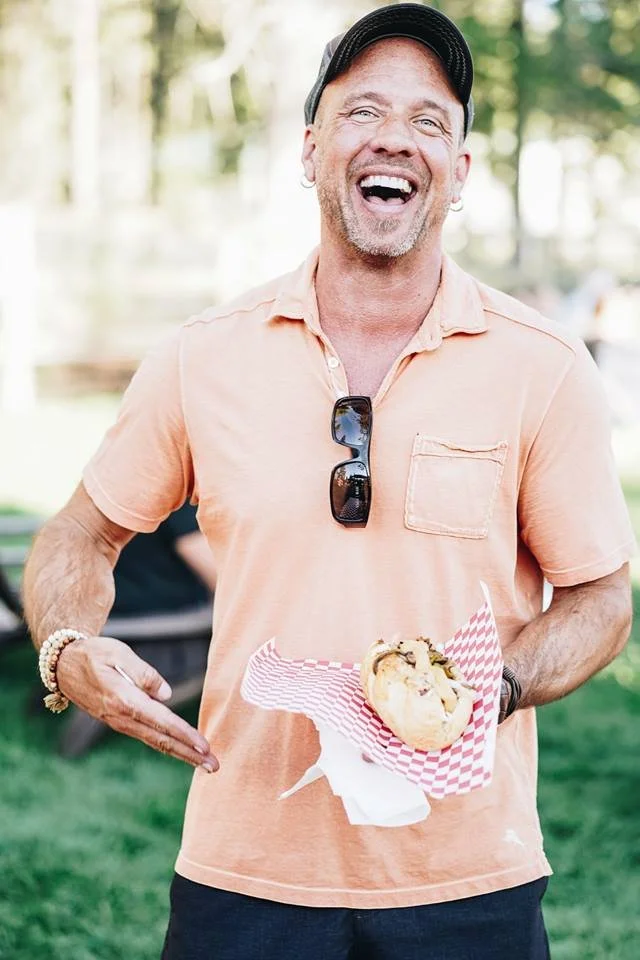 A man smiling and holding a hot dog in a checkered paper tray outdoors in a park or garden setting.