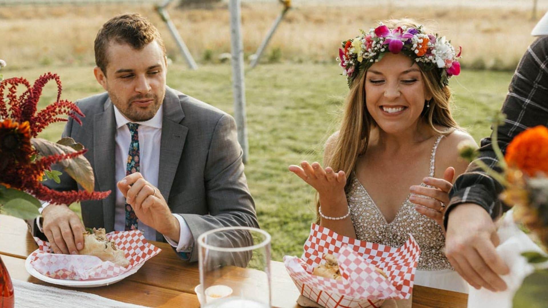 A bride wearing a flower crown and a groom in a gray suit sitting at a rustic outdoor wedding reception table enjoying food with friends and family during daytime.