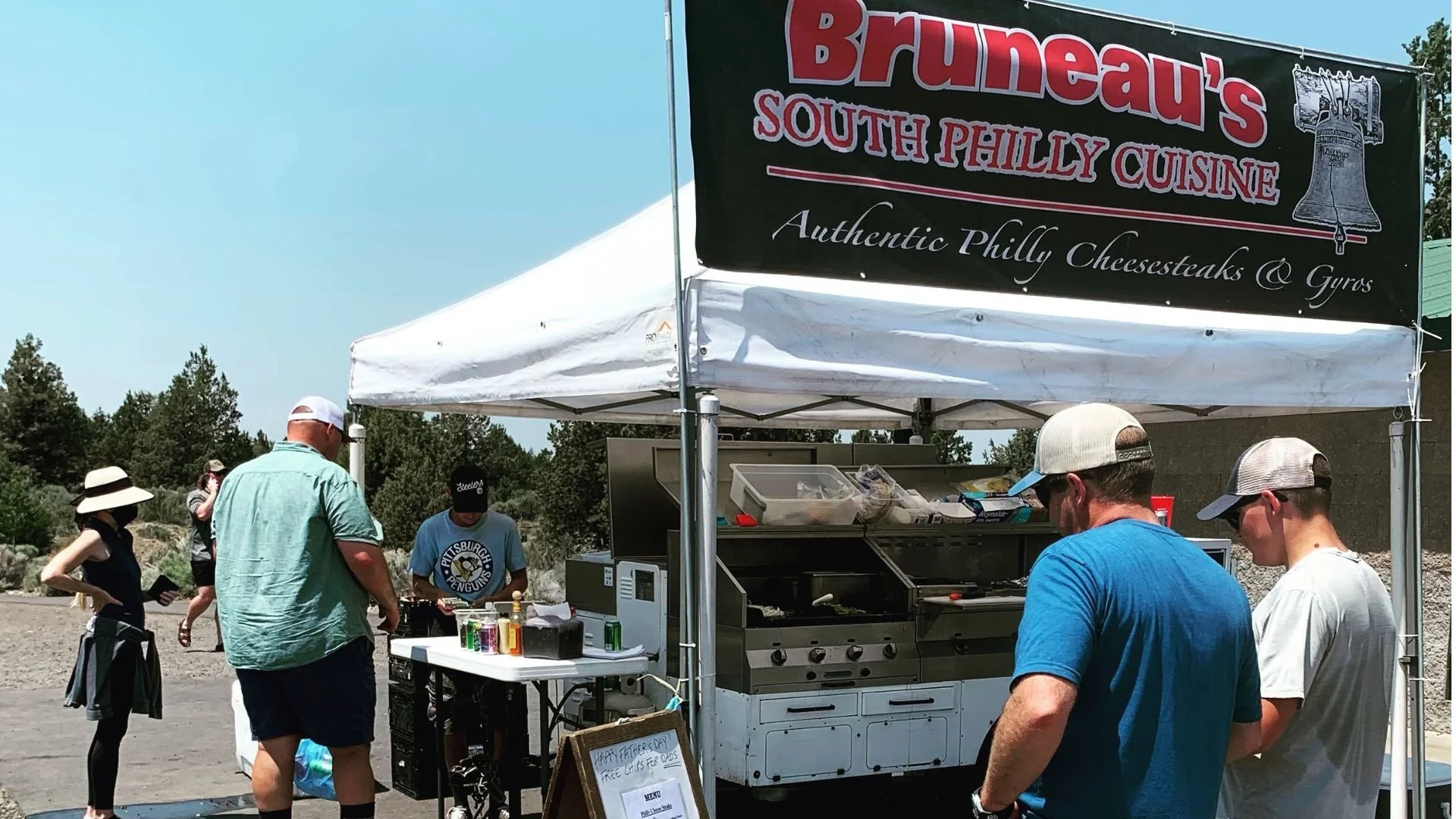 Outdoor food stand with a black and red sign that reads 'Bruneau's South Philly Cuisine: Authentic Philly Cheesesteaks & Gyros'. People are standing in line and ordering food, with a grill and condiments on a table under a white canopy.