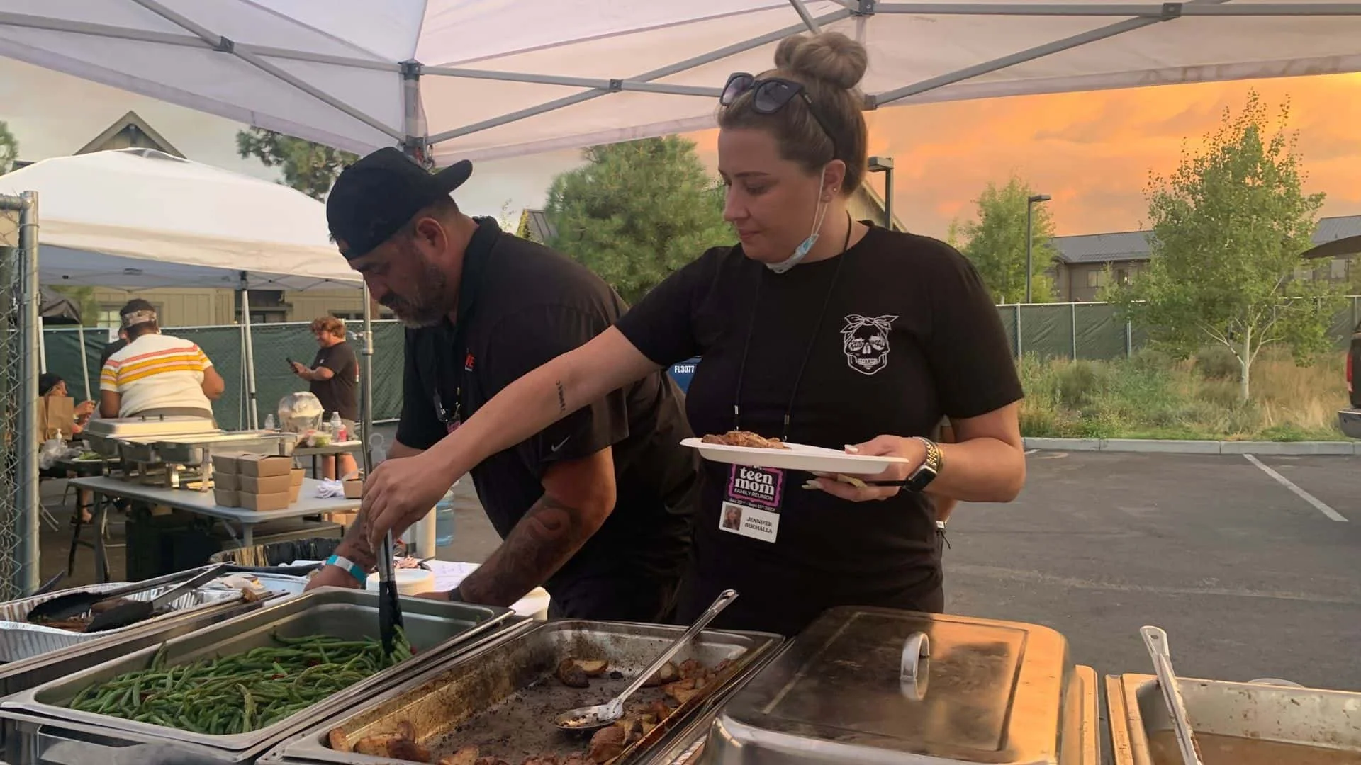 Two people serving food at an outdoor event during sunset, with a woman holding a plate and a man behind the food trays, and others in the background.