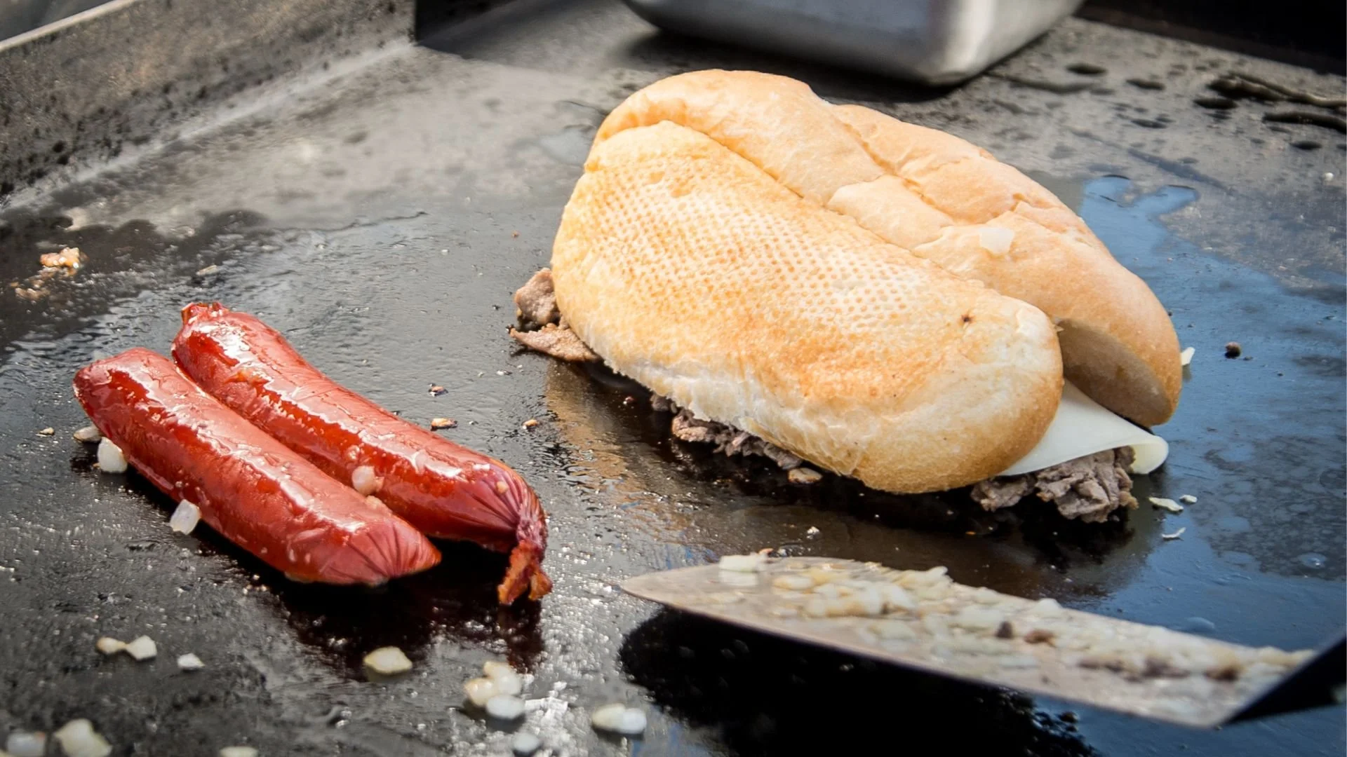Philly cheese steak and a red sausage are sizzling on a Blackstone griddle with a few scattered onions. 