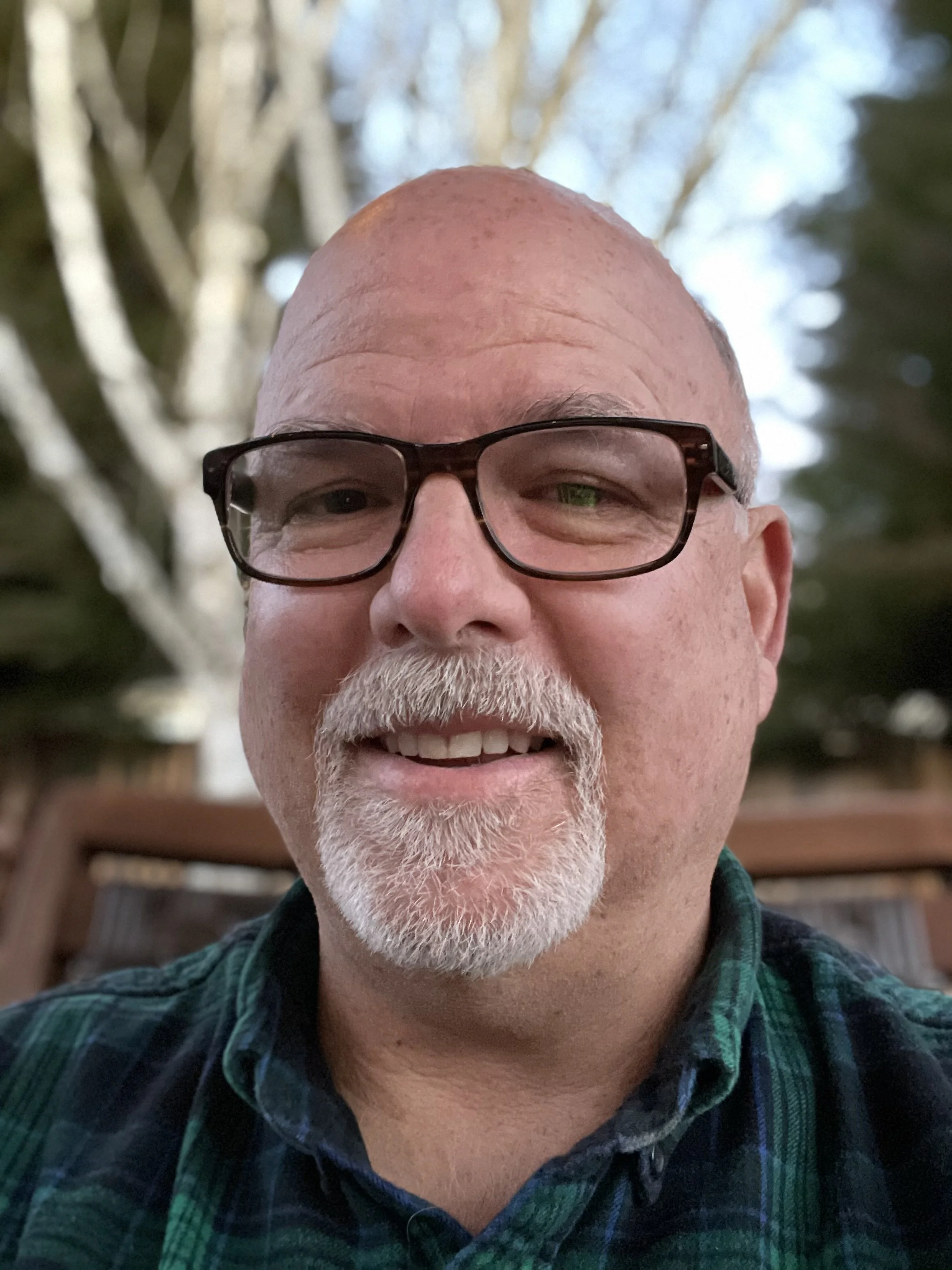 Close-up of a smiling middle-aged man with glasses, a white beard, and a bald head, outdoors in a backyard with trees in the background.