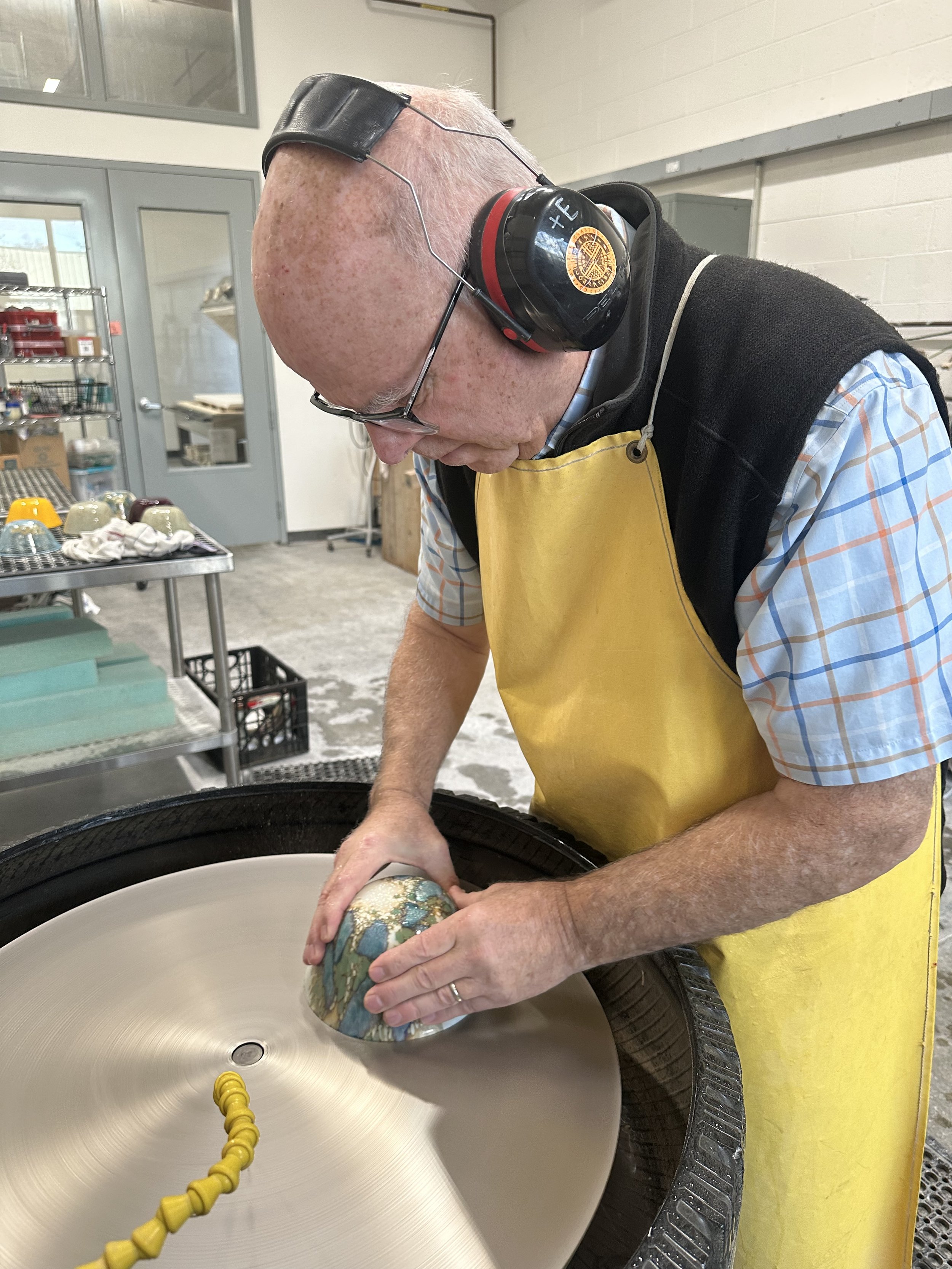 A man wearing safety headphones, glasses, a yellow apron, and a plaid shirt, is shaping a ceramic bowl on a pottery wheel in a workshop.