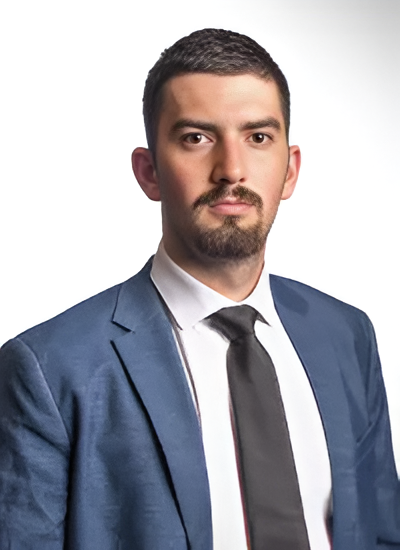 A young man with short dark hair, a beard, and mustache, dressed in a dark blue suit, white shirt, and black tie, looking serious against a plain light background.