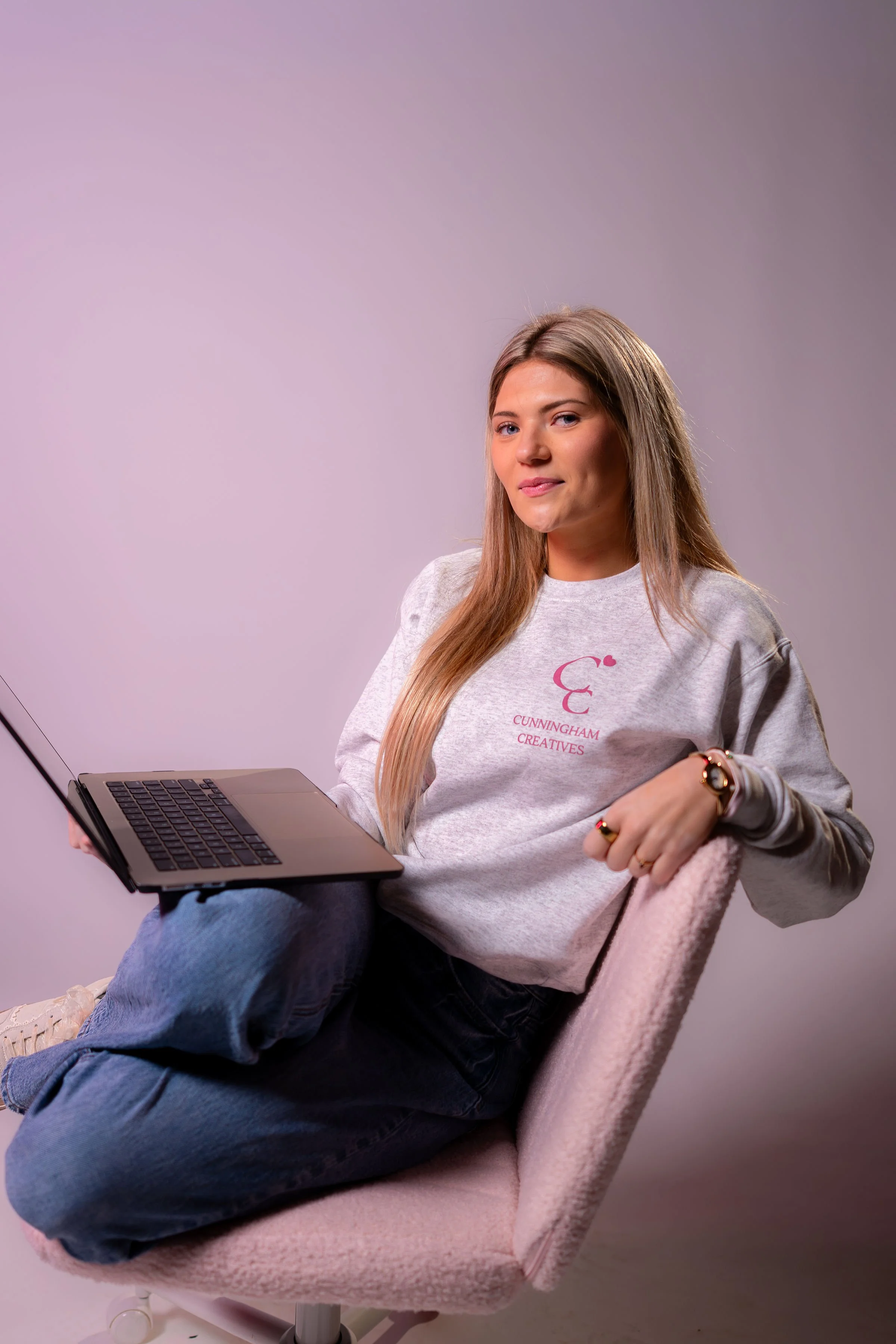 Young woman sitting on a pink chair with a laptop on her lap, wearing a gray sweatshirt with pink text that says 'Cunningham Creatives,' with a neutral expression and a plain gray background.
