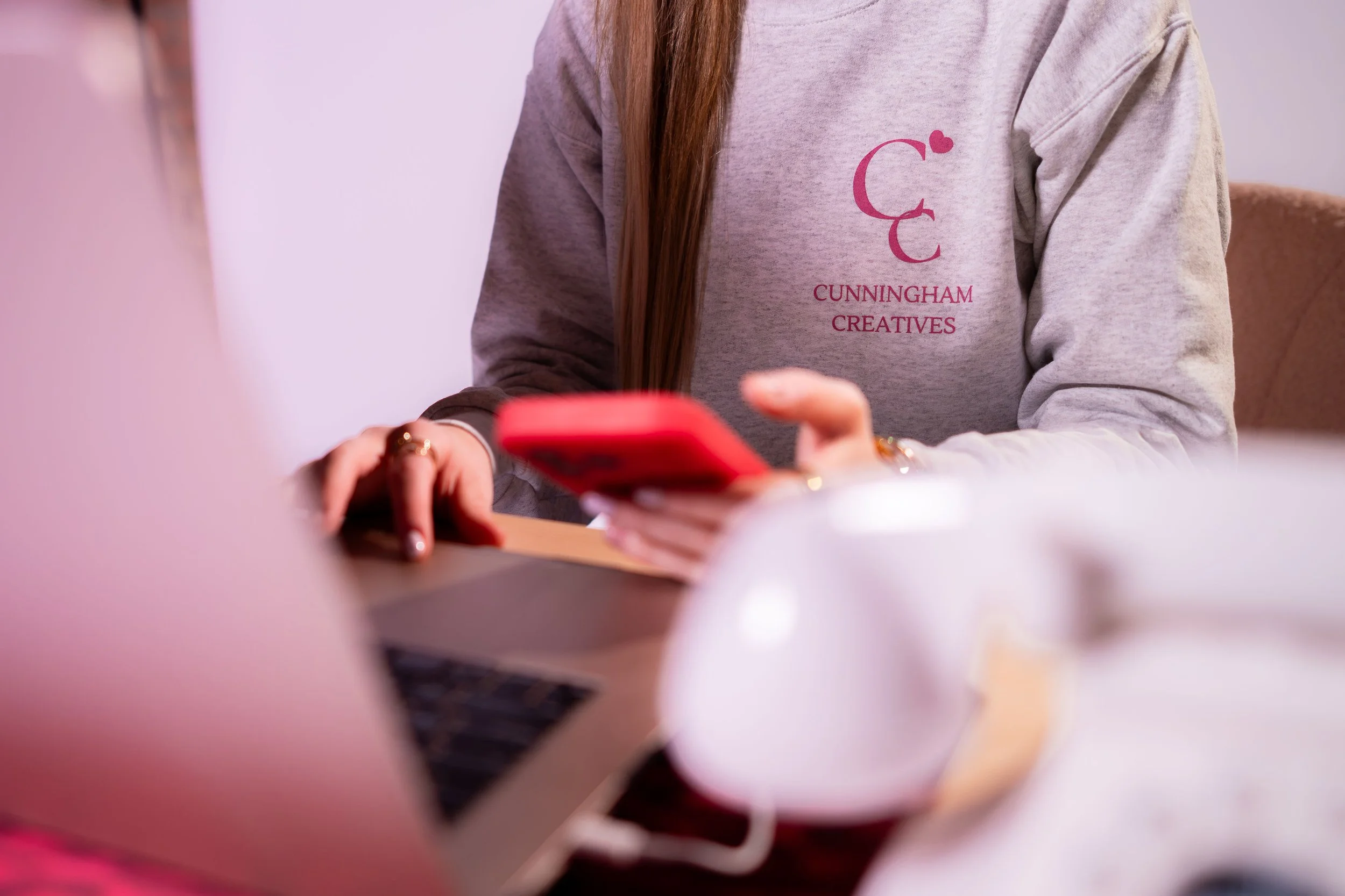 A person wearing a gray sweatshirt with the text 'Cunningham Creatives' and a logo, sitting at a desk with a laptop and using a red smartphone.