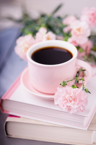 A pink coffee cup filled with black coffee on top of a stack of books, with pink flowers surrounding it.