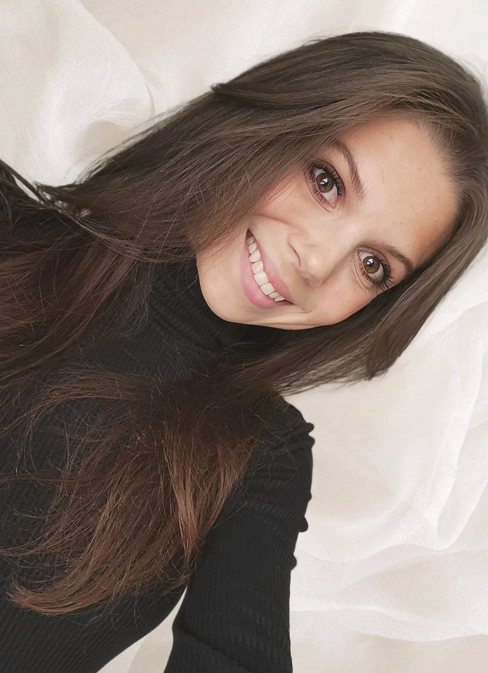 A young woman with long brown hair, smiling, wearing a black top, against a white background.