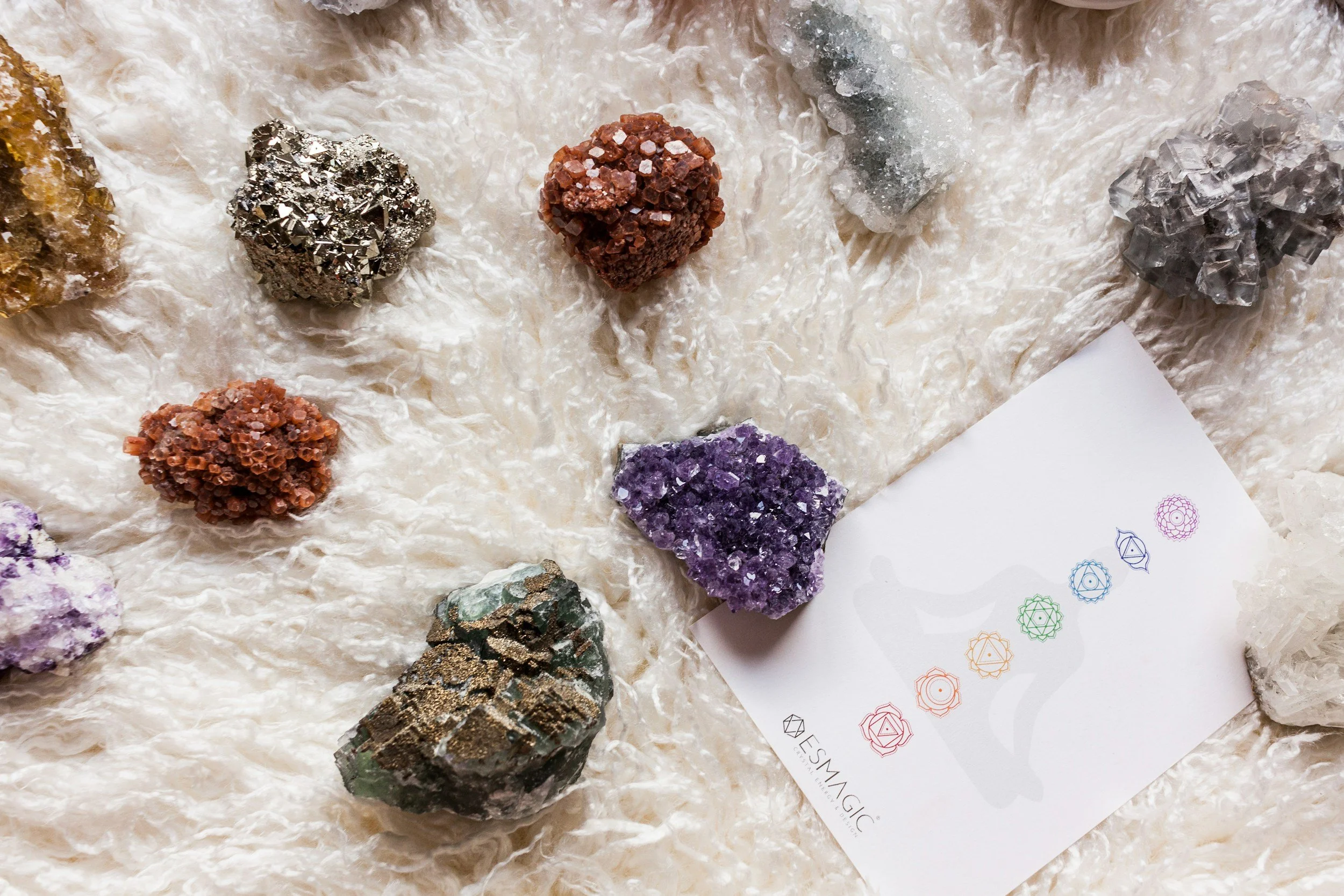 Various colorful mineral and crystal cluster specimens on a crinkled beige surface, with a white card featuring colorful geometric symbols partially visible in the lower right corner.