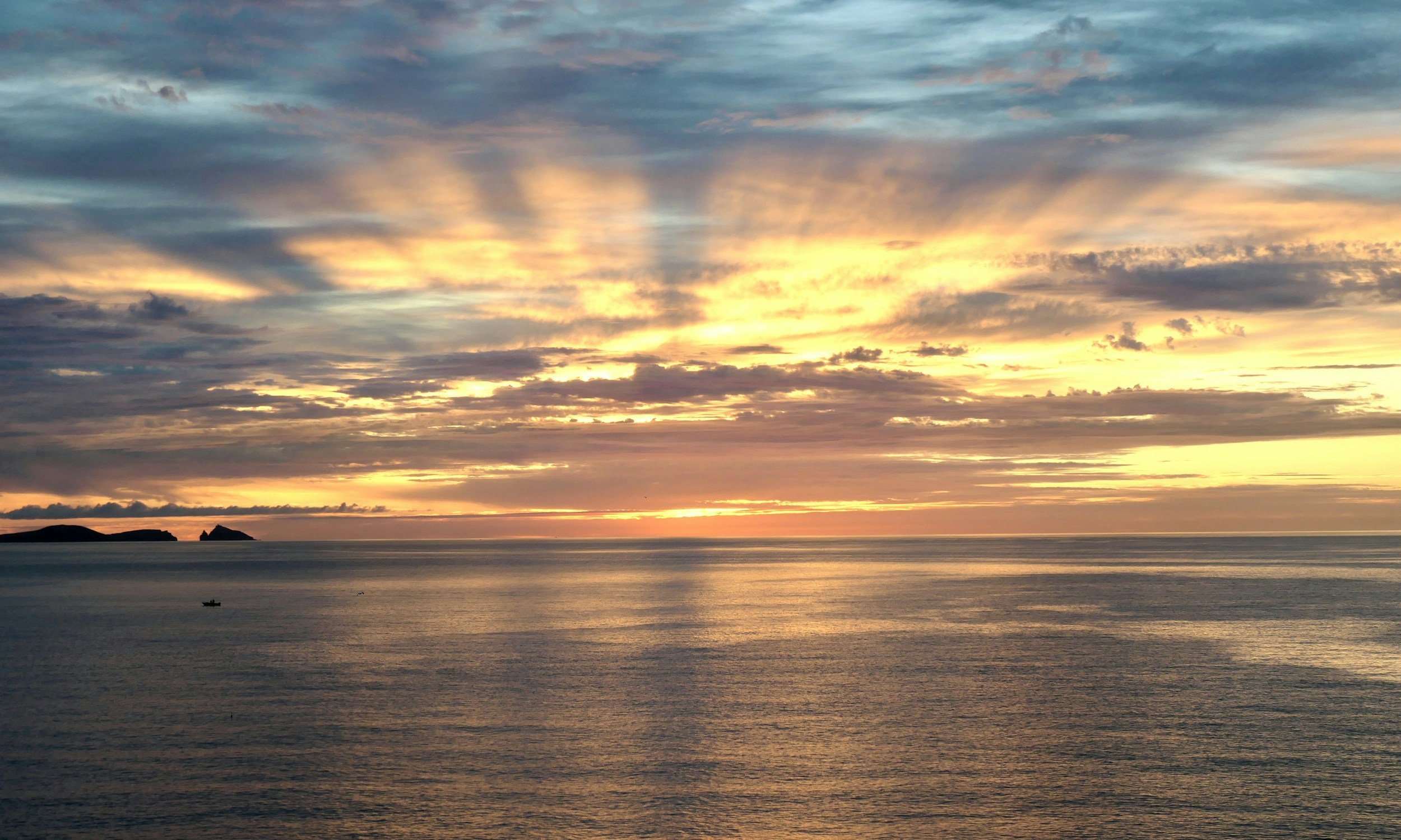 Sunset over the ocean with clouds, small islands on the horizon, and a calm water surface.