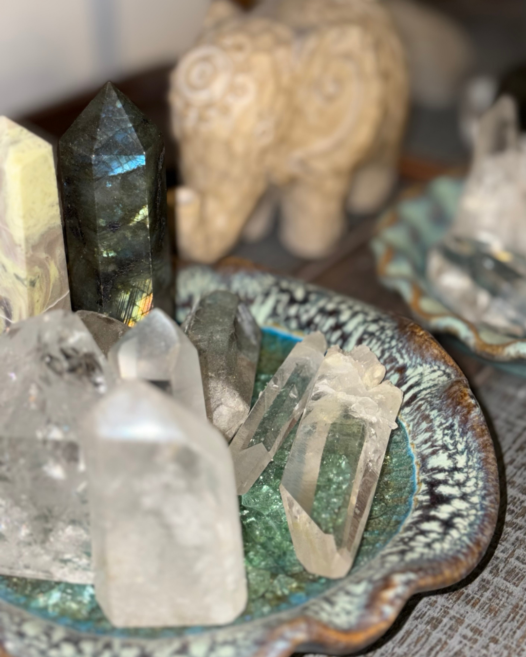 Collection of various crystals and gemstones in a decorative bowl with a carved stone sculpture in the background.