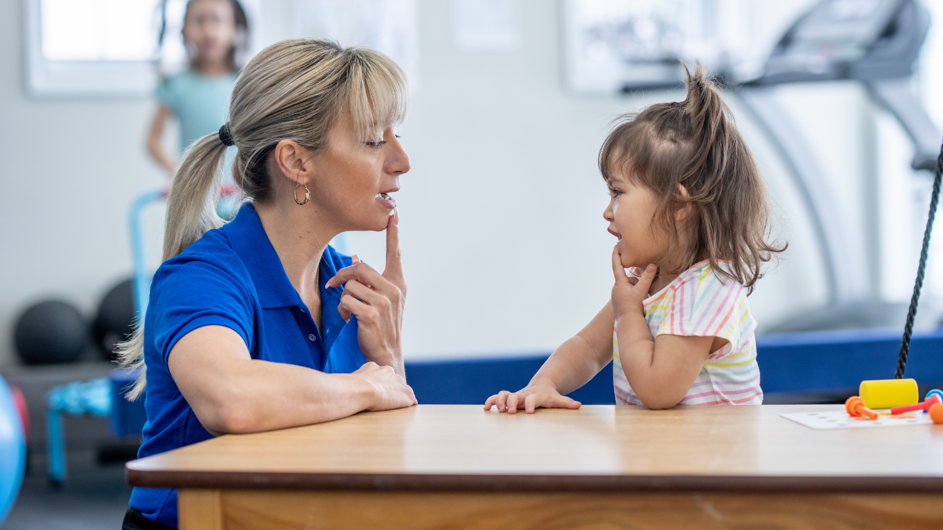A woman and a young girl face each other across a table, both with one finger on their lips, appearing to communicate quietly. The woman is wearing a blue shirt and has blonde hair tied back, while the girl is in a colorful striped shirt with brown hair.