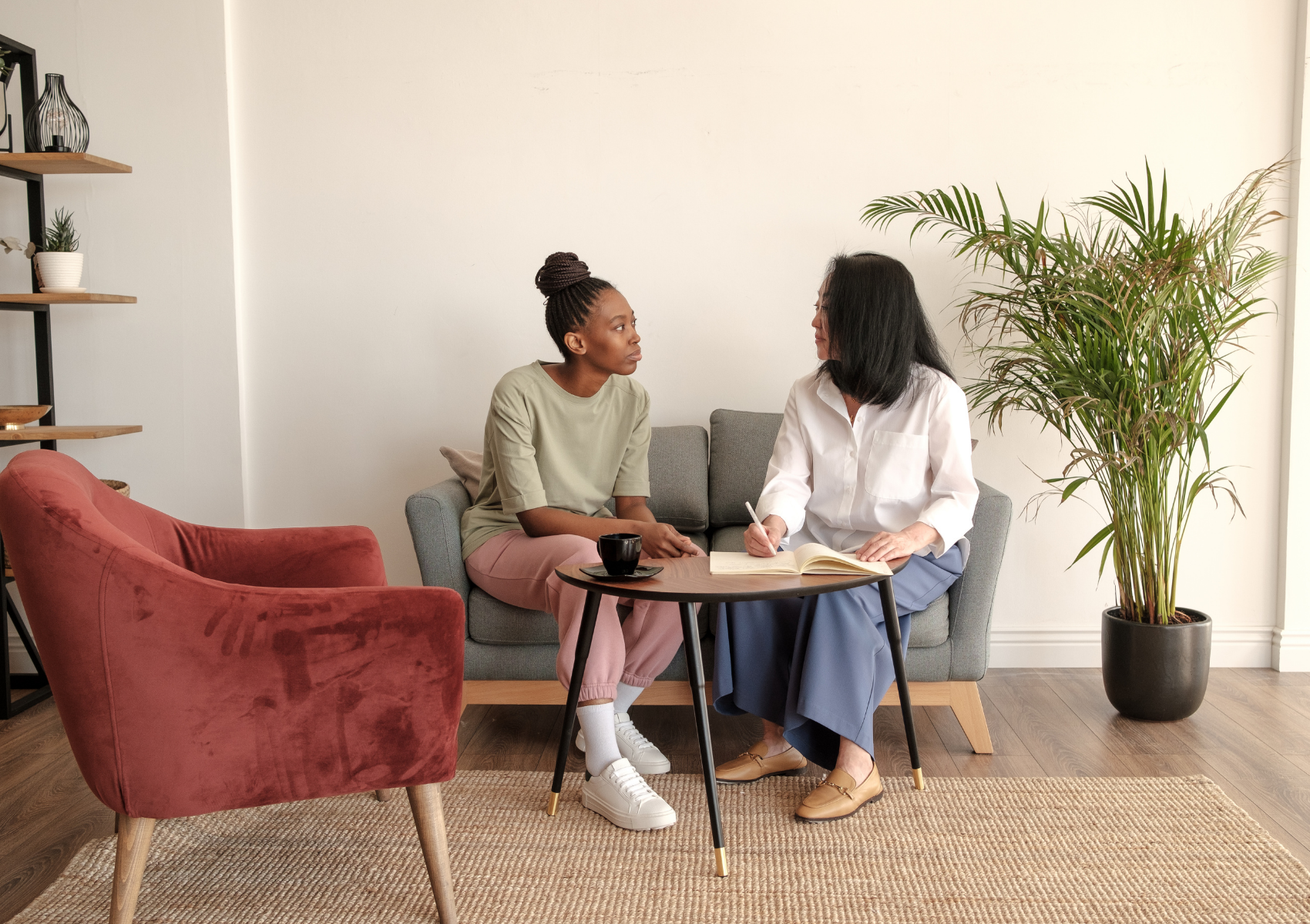Two women sit on a gray couch having a conversation with a small round table in front of them. One woman is black, wearing a green t-shirt and pink pants, the other is Asian, wearing a white shirt and blue pants. A large potted plant is beside the couch.