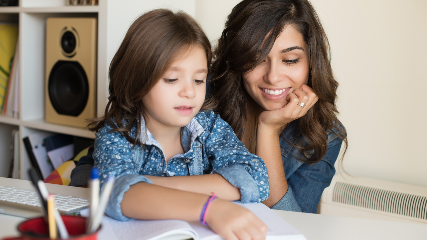 A young girl and a woman, possibly her mother, sitting at a table together. The girl is writing or drawing in a notebook, while the woman looks on with a smile, resting her chin on her hand. There are pens in a holder on the table and a bookshelf in the background.
