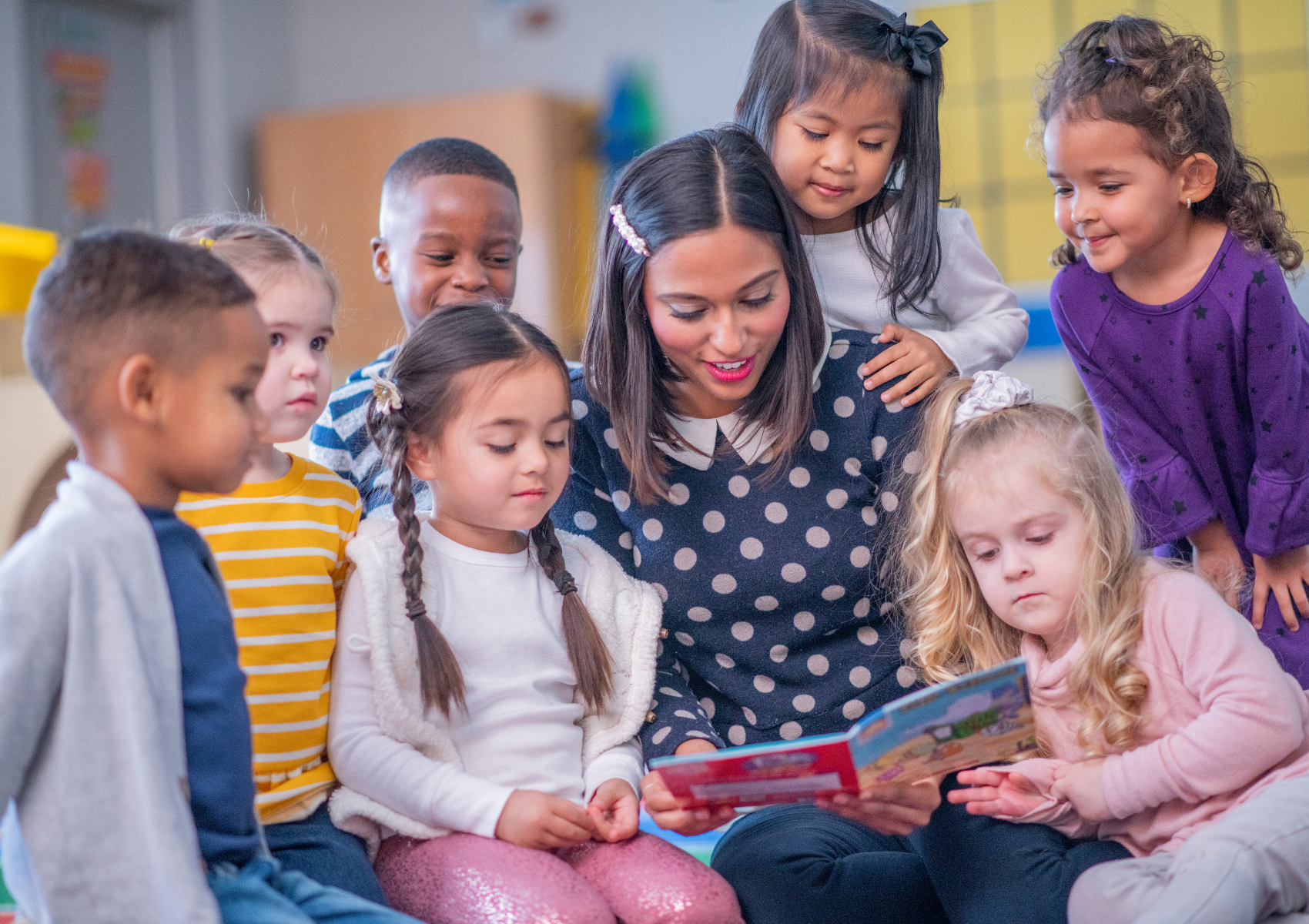 A teacher or caregiver reading a colorful picture book to a diverse group of young children in a classroom.