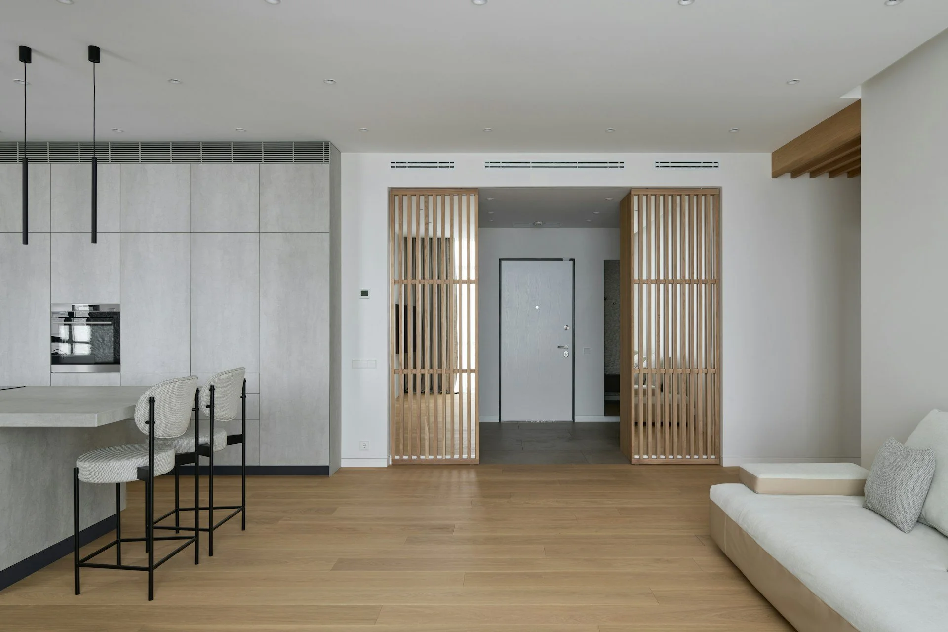 Modern living space with wooden flooring, white walls, a beige sofa, a kitchen area with light-colored cabinets, and wooden slatted room dividers leading to an entryway.