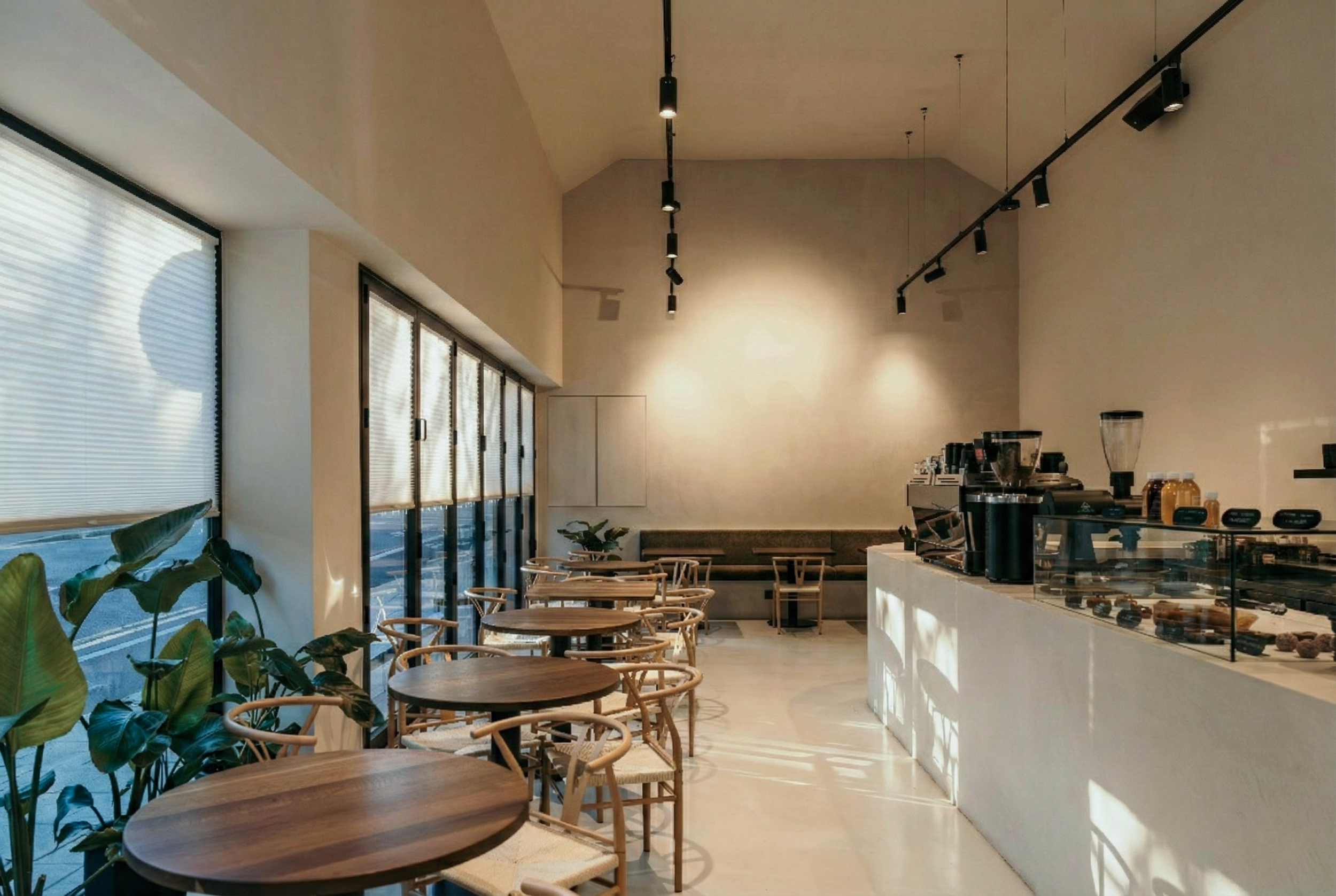 Empty cafe interior with wooden tables and chairs, large windows with blinds, plants near the window, and a white counter with coffee equipment and pastries.