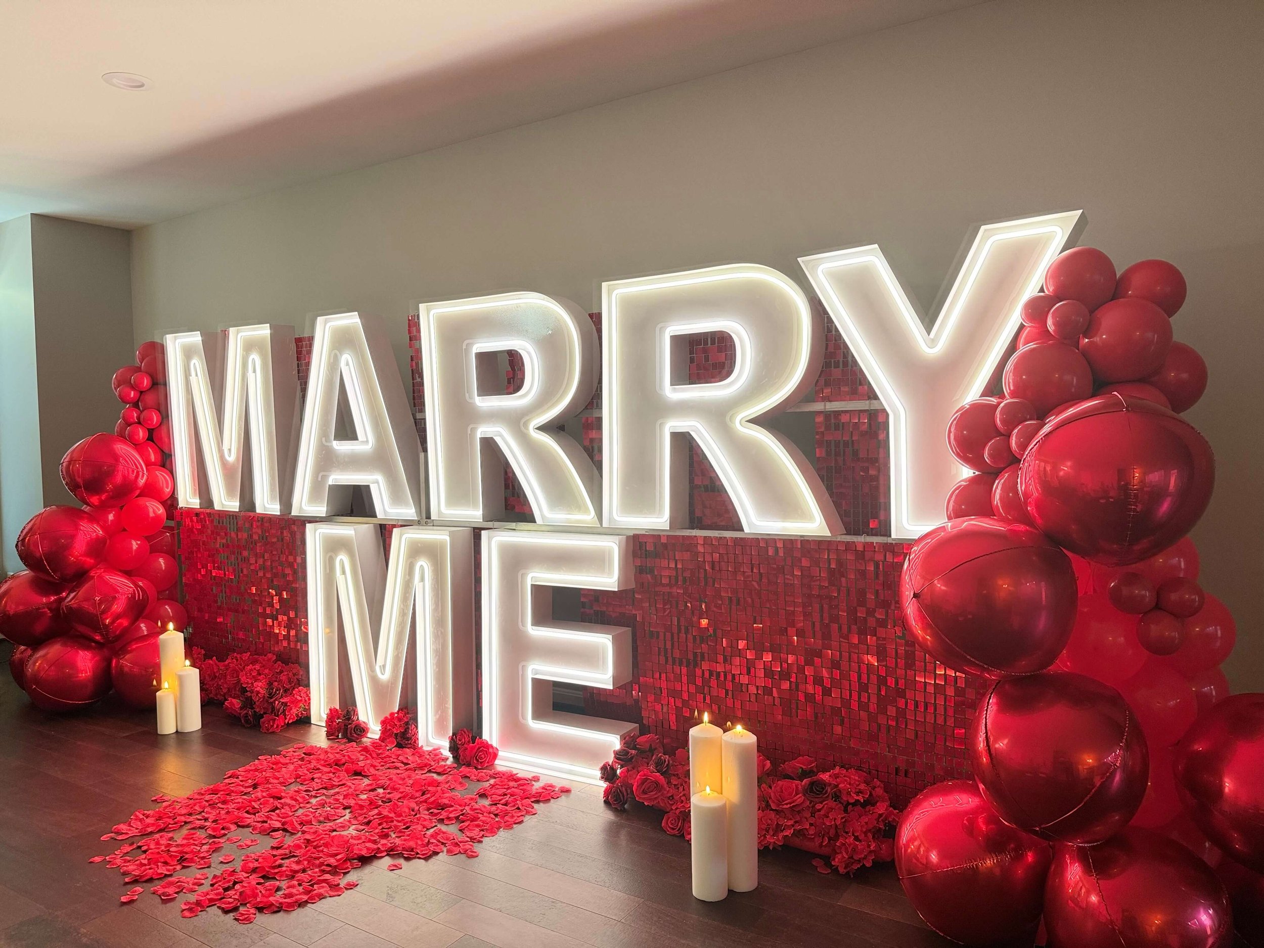 Light-up LED marquee letters spelling “MARRY ME” with red balloons, candles, and rose petals, styled for a romantic proposal event.